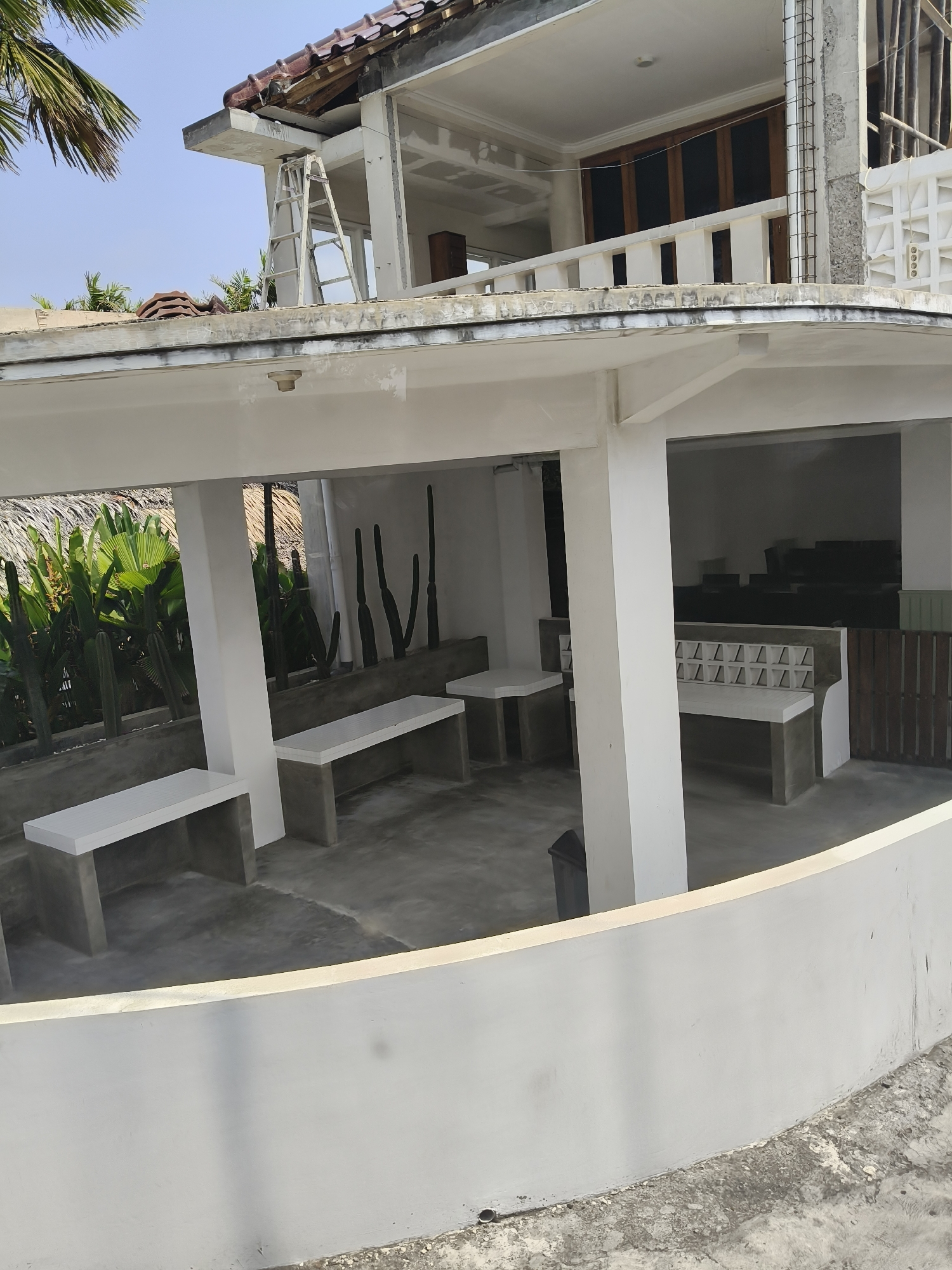 The image depicts a partially constructed building in Desa Batukaras, Indonesia. The foreground features a curved, white, low wall. Beyond the wall, an open-air space is visible, with concrete flooring and several white, rectangular benches supported by concrete bases. Two of these benches are prominently placed on the left and in the center of the frame. To the right, another long, white bench with a decorative backrest of interlocking squares is situated.

In the background, the building appears to be undergoing renovation or construction. A section of the upper floor is incomplete, with exposed concrete columns and rebar. A ladder leans against the structure. Various architectural elements, including wooden-framed windows and a tiled roof, are visible. Lush green foliage, including palm leaves and cacti, surrounds the building, suggesting a tropical or sub-tropical environment. The sky above is clear and blue. The overall scene conveys a sense of ongoing development and an outdoor seating area.