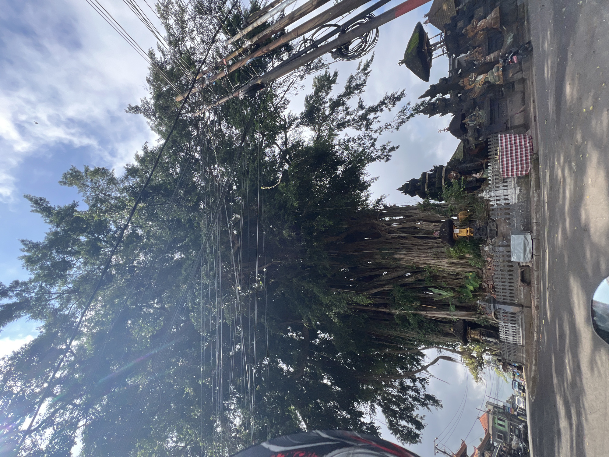 The image shows a large banyan tree with extensive aerial roots in Tembuku, Indonesia. The tree is situated in front of a complex of stone structures, which appear to be religious or decorative, featuring tiered roofs and carved figures. A grey metal fence with ornate detailing separates the tree and structures from the paved road. Power lines are strung across the upper part of the image, passing through the branches of the tree. To the left, buildings with orange roofs and a street with several parked vehicles are visible, indicating an urban or semi-urban setting. The sky is partly cloudy with bright sunlight filtering through the leaves. The overall scene suggests a cultural or historical site within a lively Indonesian town.