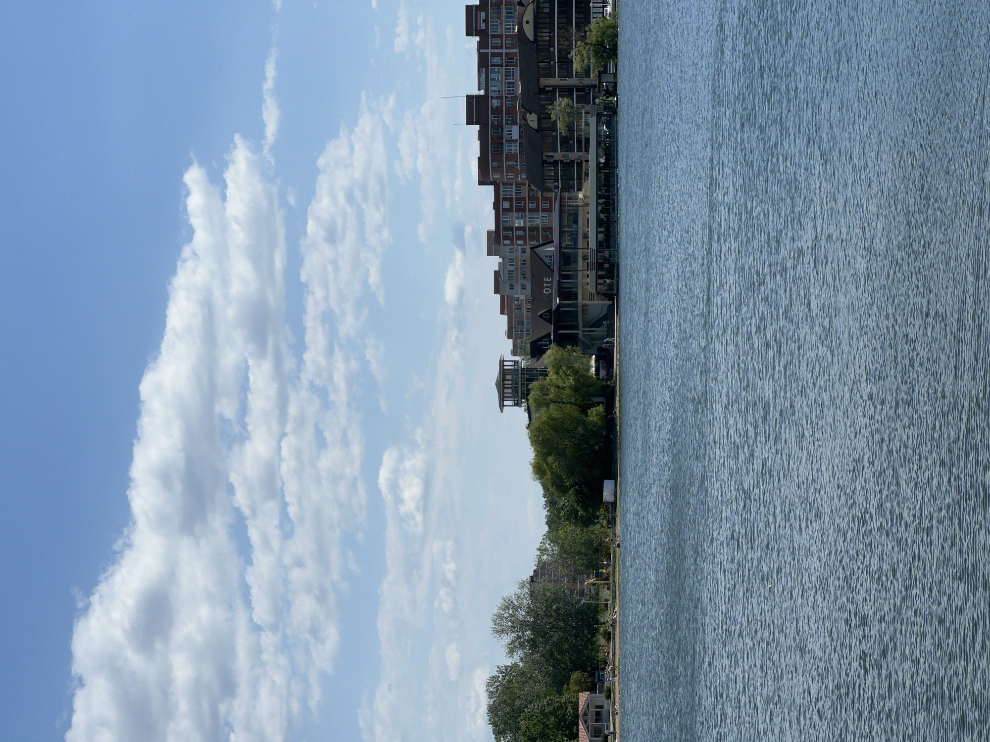 The image captures a cityscape alongside a body of water, likely a river or lake. The sky is partially covered with cumulus clouds, indicating fair weather. On the left of the image, several buildings of varying heights can be seen, some appearing to be residential or commercial structures. A line of trees runs along the shoreline. The water surface shows slight ripples.
Without additional reference points in the image, definitively placing this scene within Goryachiy Klyuch, Russia is impossible.