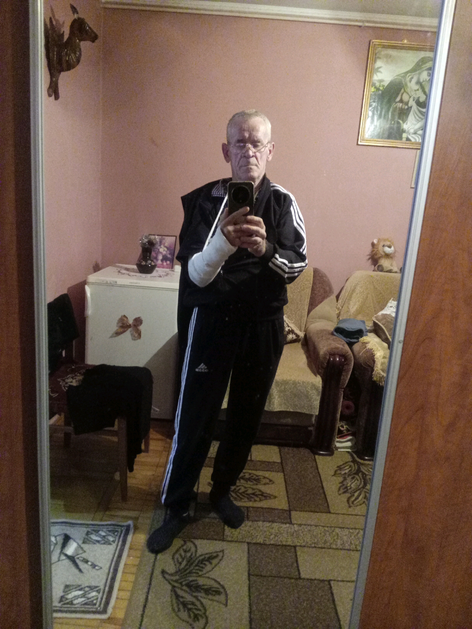 A man, appearing to be in his 50s or 60s, with short gray hair and glasses, is taking a selfie in a mirror. He is wearing a black tracksuit with white stripes on the sleeves and pants, and has his right arm in a cast. The man is standing in what appears to be a living room or bedroom, with a dresser or refrigerator to his left, and a sofa with a stuffed lion on it to his right. The walls are painted pink, and there are several framed pictures and decorative items on the walls and furniture. The floor is covered with a patterned rug. The man is standing on a rug with a geometric pattern. The overall impression is of a modest, somewhat cluttered living space. There is no indication of the specific location within Tiachiv, Ukraine.