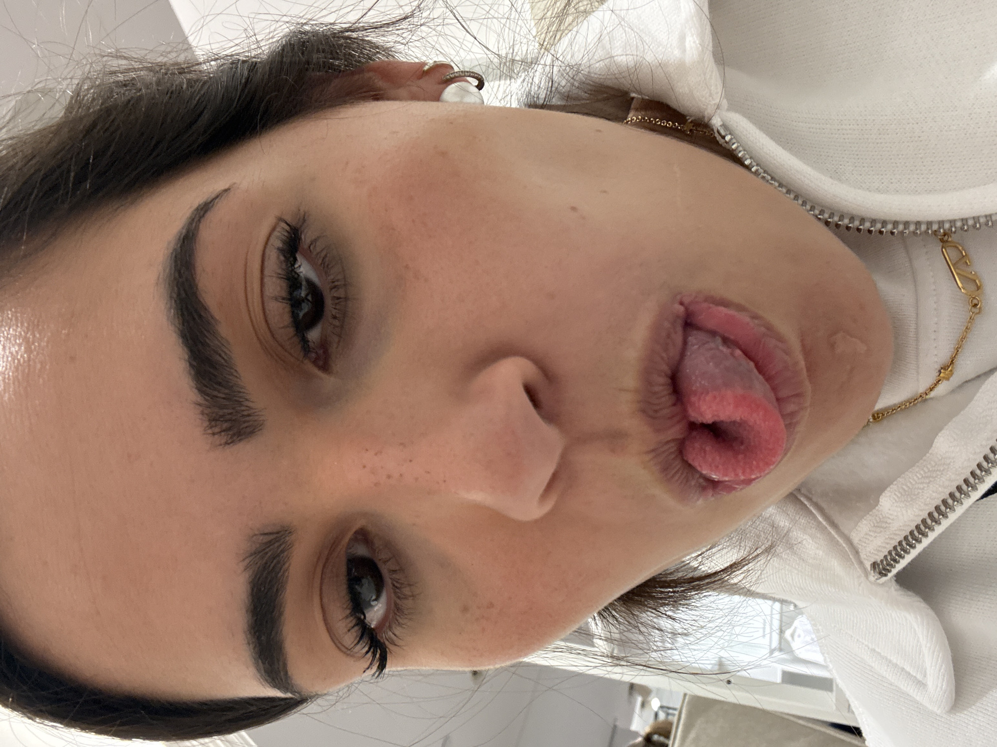 A close-up, tilted, eye-level shot depicts a woman with dark eyebrows and eyelashes, light skin with freckles, and dark hair. She is sticking out her tongue, which is rolled and has a texture of small bumps. She is wearing a white hoodie with a gold chain necklace featuring a 'V' pendant. An earring is visible in her ear. The location is not determinable from the image.