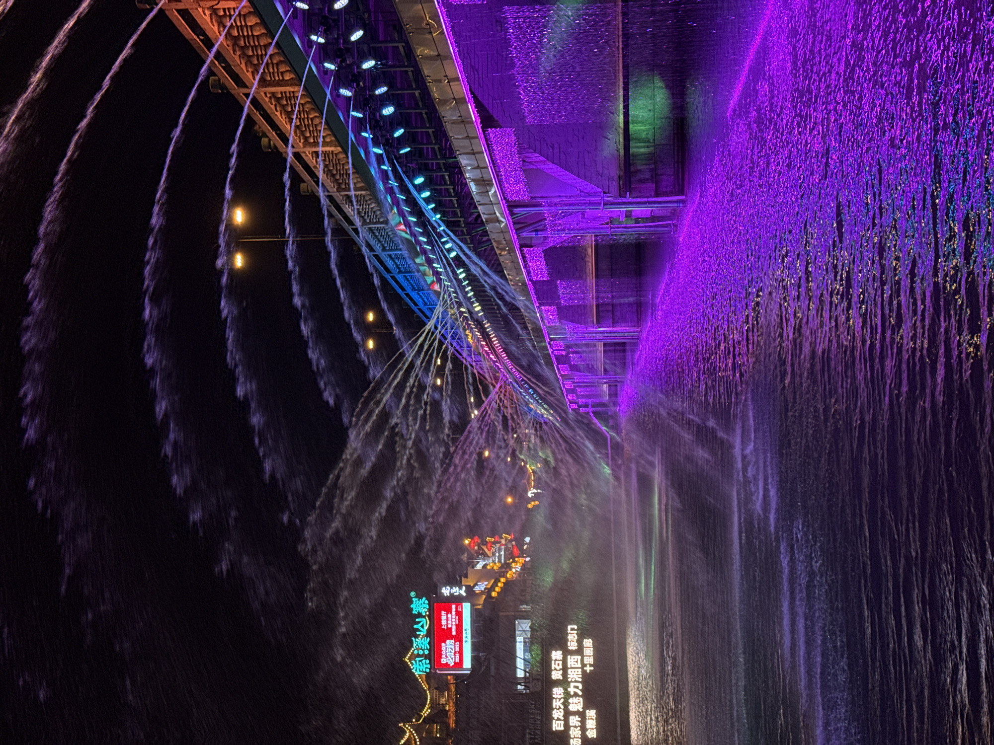 The image depicts a night scene in Wulingyuan, China, featuring an illuminated bridge with water features. The bridge is adorned with multicolored lights, including purple, blue, and orange hues. Water jets arc away from the bridge, creating a dynamic display. A dense curtain of water falls in the foreground, illuminated in purple. A sign displaying Chinese characters and the words "大众点评 上榜餐厅", indicates a featured restaurant. Additional signage includes "百龙天梯 黄石寨" and "汤家界 魅力湘西". The overall environment is dark, emphasizing the colorful illumination of the bridge and water features.