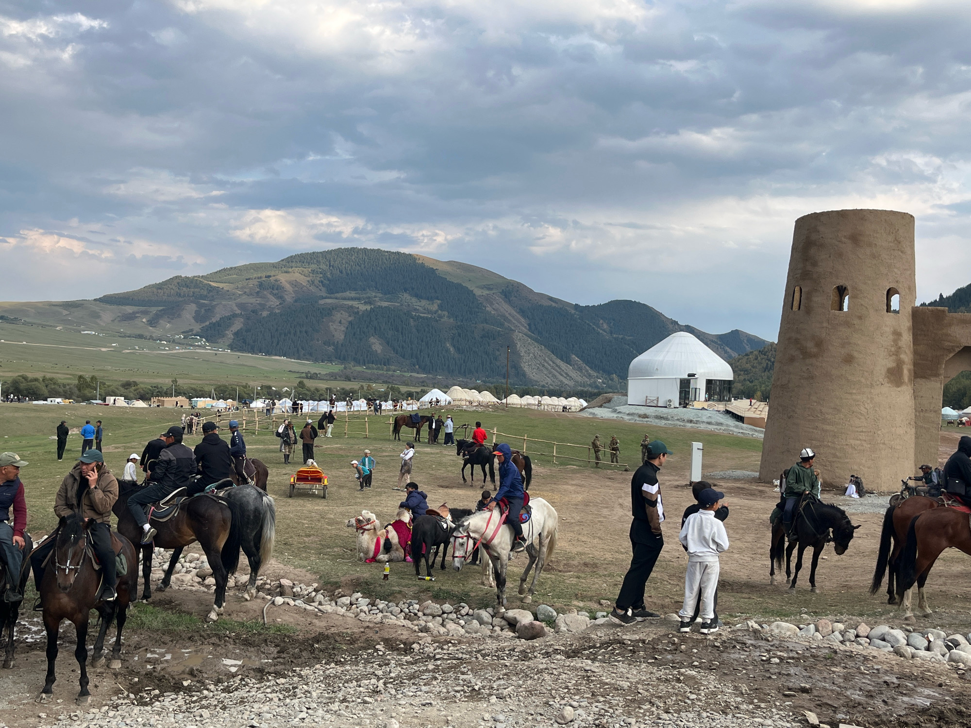 The image depicts a scene in Grigor'yevka, Kyrgyzstan, likely at a cultural event or festival. In the foreground, several individuals are on horseback, with some people walking around them. A person is on the phone while riding a horse. Two camels are present, one of which is lying down. A small red cart is also visible. In the background, there are yurts and a large white dome structure, possibly a tent or pavilion. A distinctive, tall, rough-textured, cylindrical tower with arched openings stands prominently on the right side of the frame. Rolling, tree-covered hills form the backdrop under a cloudy sky.