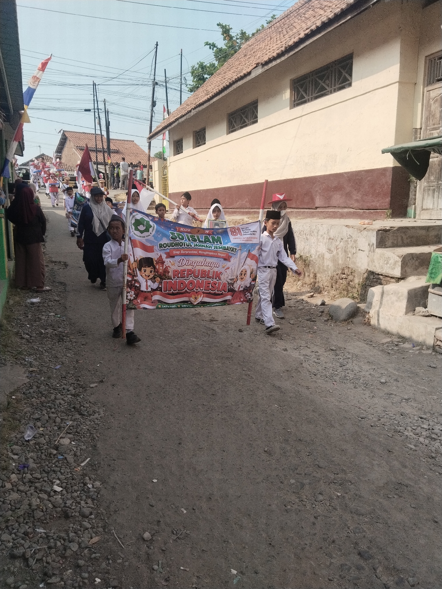 In Jembayat, Indonesia, a group of children participate in a parade. The children, dressed in white uniforms and head coverings, walk along a dirt road lined with simple buildings. They hold flags and a banner that reads "SD Islam Roudhotul Jannah Jembayat" and "Dirgahayu Republik Indonesia." The parade appears to be a celebration of Indonesian Independence Day. The buildings are one or two stories high, with tiled roofs and cream-colored walls. Electrical wires crisscross overhead, and trees are visible in the background. The road is unpaved, with gravel and dirt. Some of the children are smiling and waving. The overall scene suggests a festive and community-oriented event.