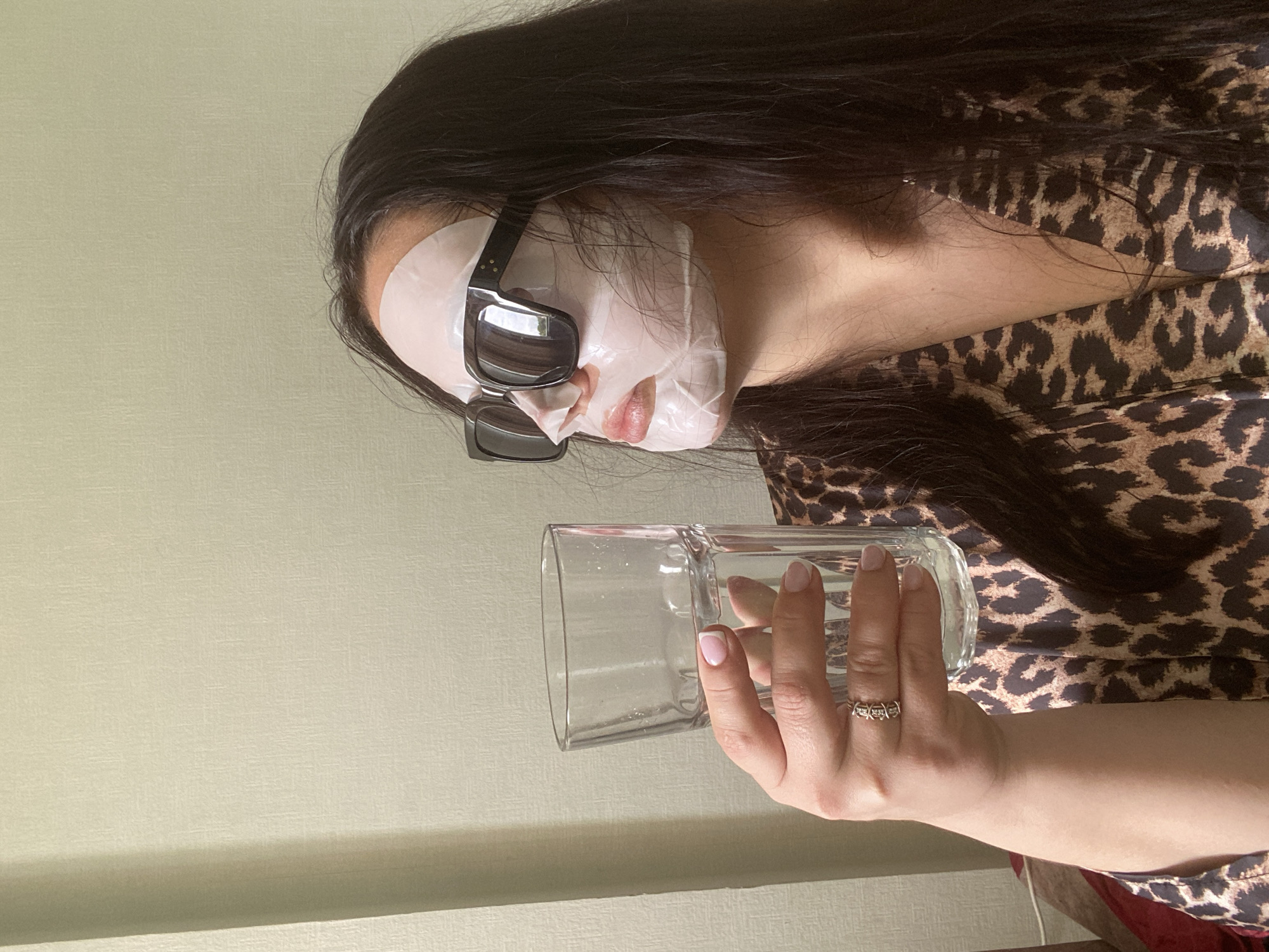 A woman with dark hair, wearing a face mask and sunglasses, is holding a glass of water. She is wearing a leopard print robe or top. The background appears to be a light-colored wall. There is no visual information to indicate the location is in Sochi, Russia.