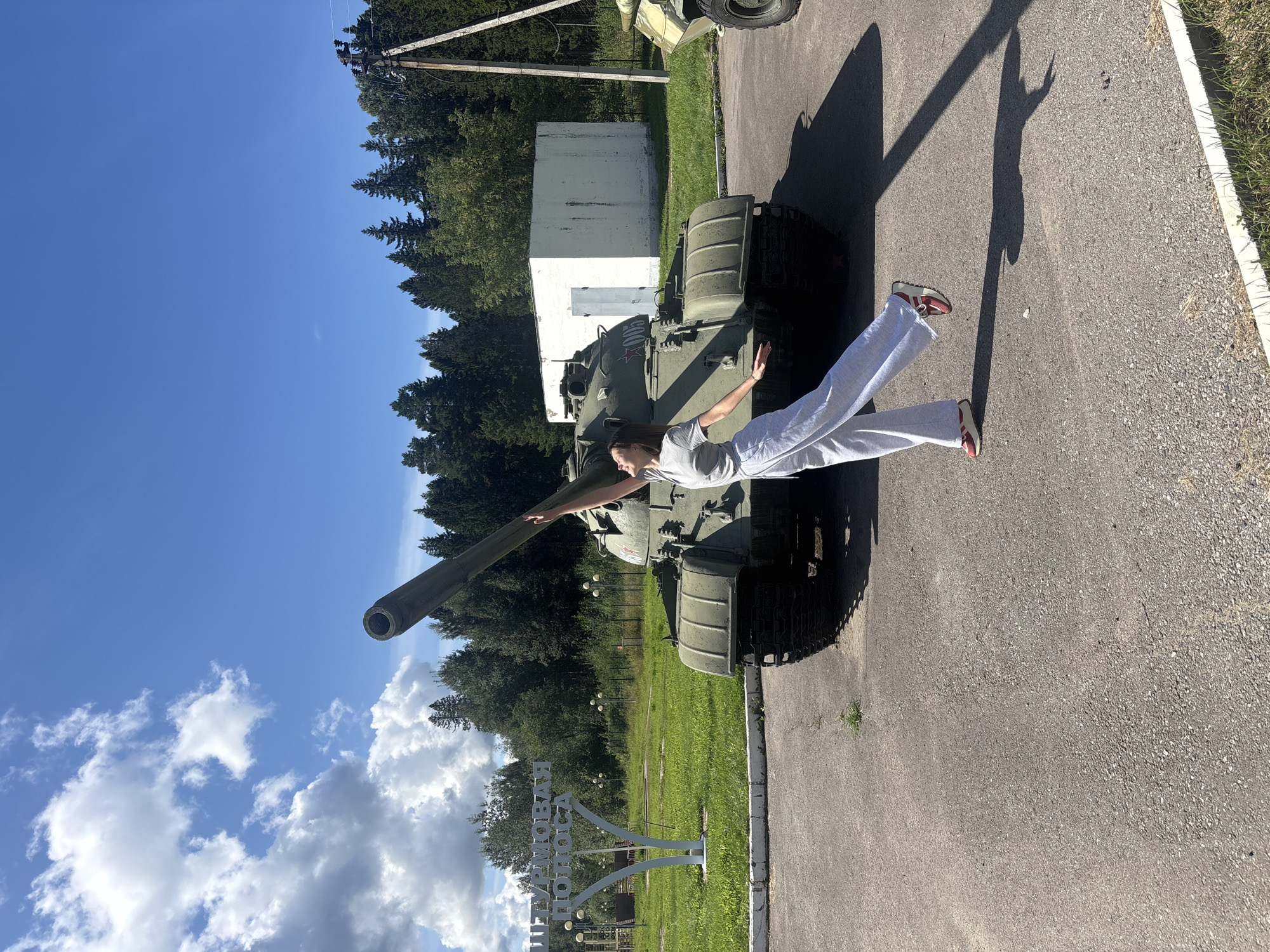 A young woman stands in front of a Soviet-era tank in the city of Gamy, Russia. The tank, painted olive drab, is parked on an asphalt surface in front of a white building and a treeline. The woman, wearing a grey t-shirt and light grey wide-leg pants, is reaching up towards the tank's barrel, her body angled in a dynamic pose. Her left leg is extended, and she appears to be in mid-movement. Behind the tank, a sign with Cyrillic lettering reads "ШТУРМОВАЯ ПОЛОСА" which translates to "Assault Course". The sky is a clear blue with scattered white clouds. The scene suggests an outdoor exhibit or memorial.