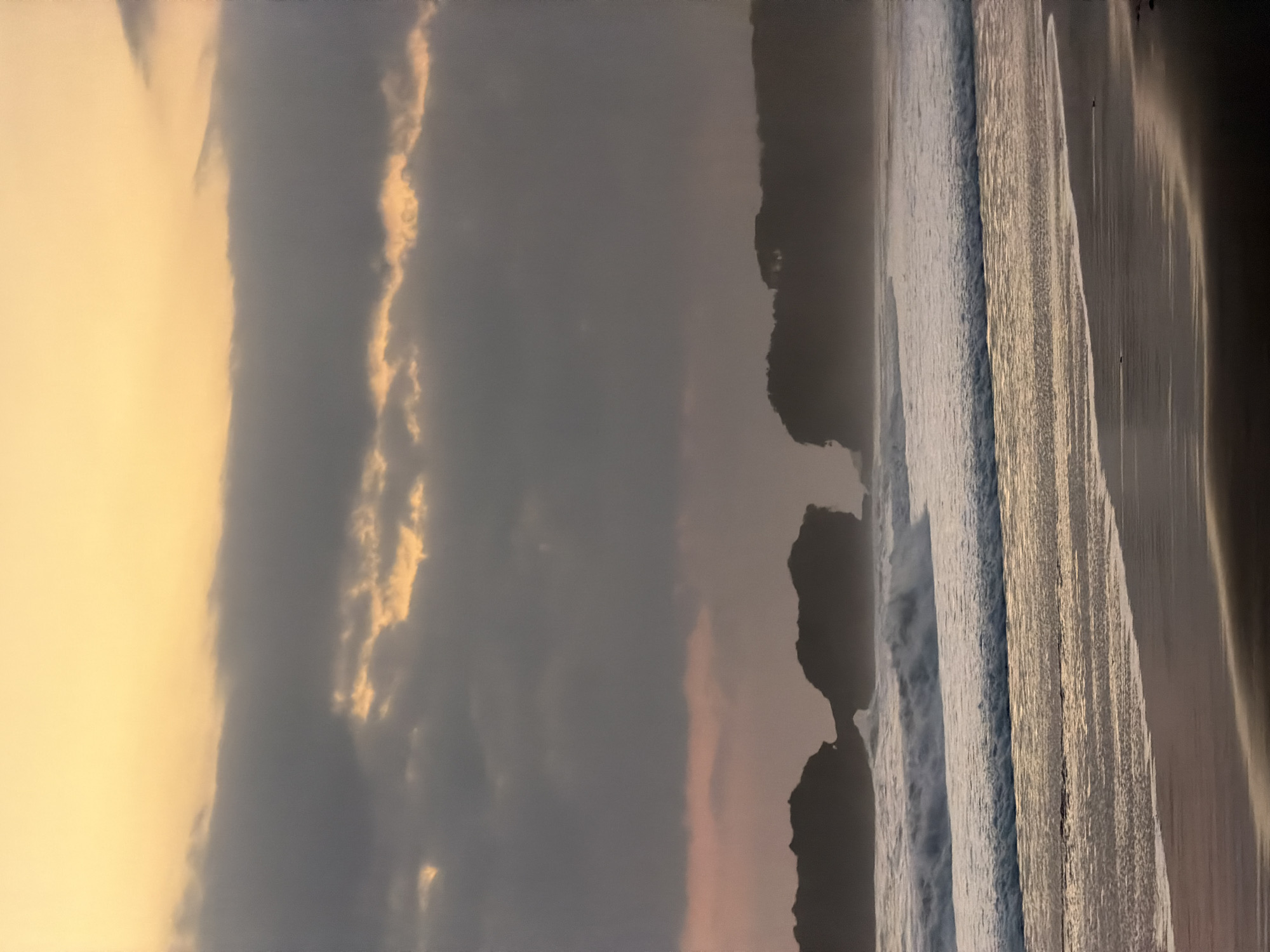 This image captures a wide coastal landscape in Selemadeg Timur, Indonesia, at either sunrise or sunset. The upper portion of the sky is clear and bright yellow-orange, transitioning downwards into a broad, horizontal band of dark grey clouds. Within this cloud layer, streaks and patches of the brighter yellow-orange light are visible, indicating breaks in the cloud cover. Below the main cloud mass, the sky lightens to a soft, desaturated pinkish-grey at the horizon.
Along the horizon, dark, silhouetted landforms emerge from the ocean. On the left, two large, rugged rock formations are present, appearing close together with a narrow gap or arch between them. To the right, a more extensive, dark landmass, likely a vegetated cliff or headland, extends into the scene, its details obscured by shadow and distance.
The ocean occupies the mid-ground, displaying multiple lines of waves breaking and rolling towards the shore. The most prominent wave, positioned centrally, exhibits a distinct white foam crest. Closer to the foreground, shallower waves extend across the frame. The wet sand of the beach in the foreground reflects the soft light, appearing dark brown with subtle golden and pinkish tones, and features horizontal lines left by the receding tide. The beach surface is smooth and uninterrupted. No people, animals, or man-made structures are discernible within the visible area.