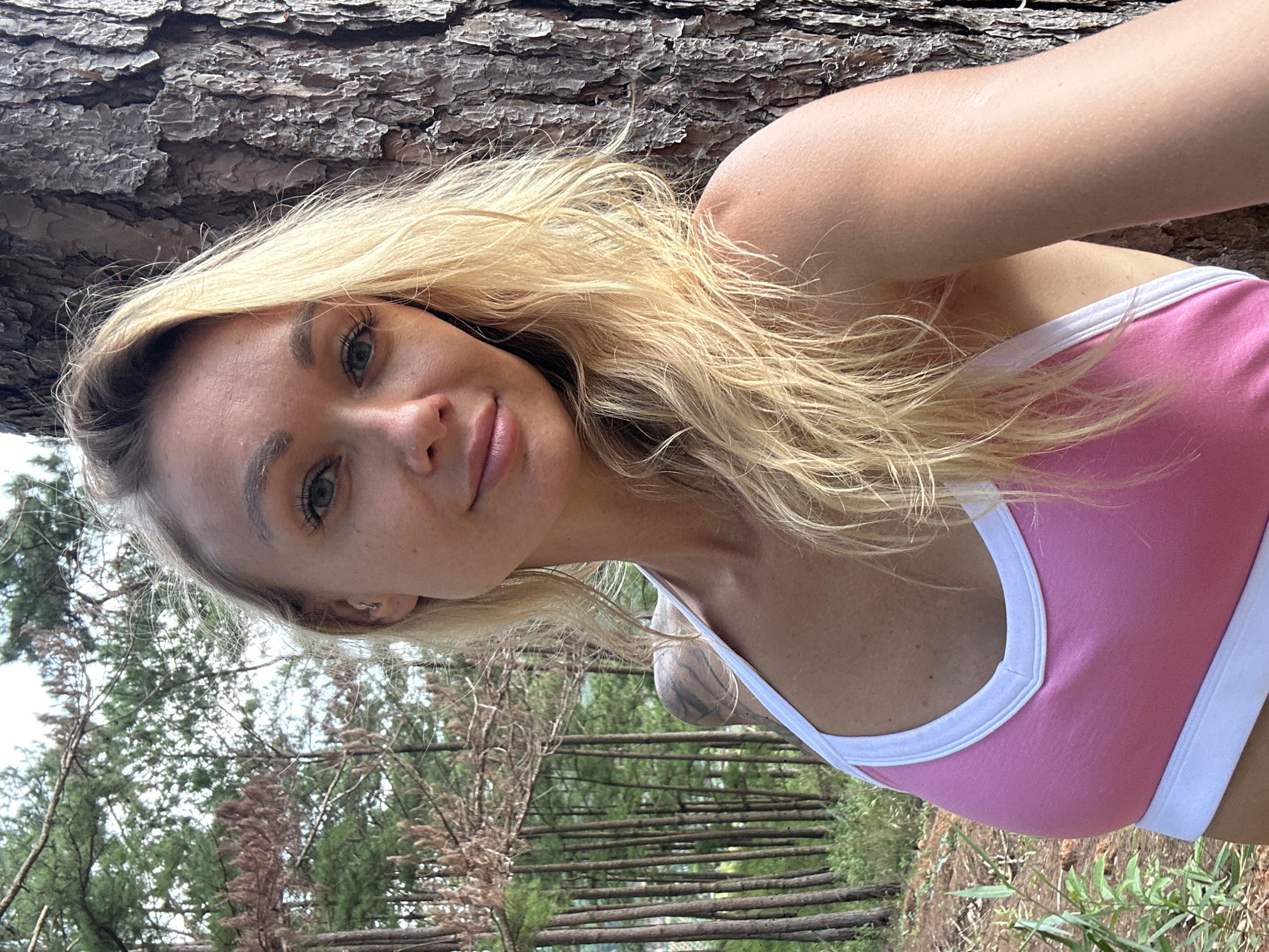 A blonde woman with blue eyes and light skin is shown in a close-up selfie. She is wearing a pink and white tank top and leaning against a tree. Her hair is long and blonde, framing her face. The background consists of a forest with tall, slender trees and some greenery. The lighting suggests it is daytime. The image appears to have been taken outdoors, possibly in a natural setting. There are no discernible urban elements to suggest it is in Thôn 3, Vietnam.