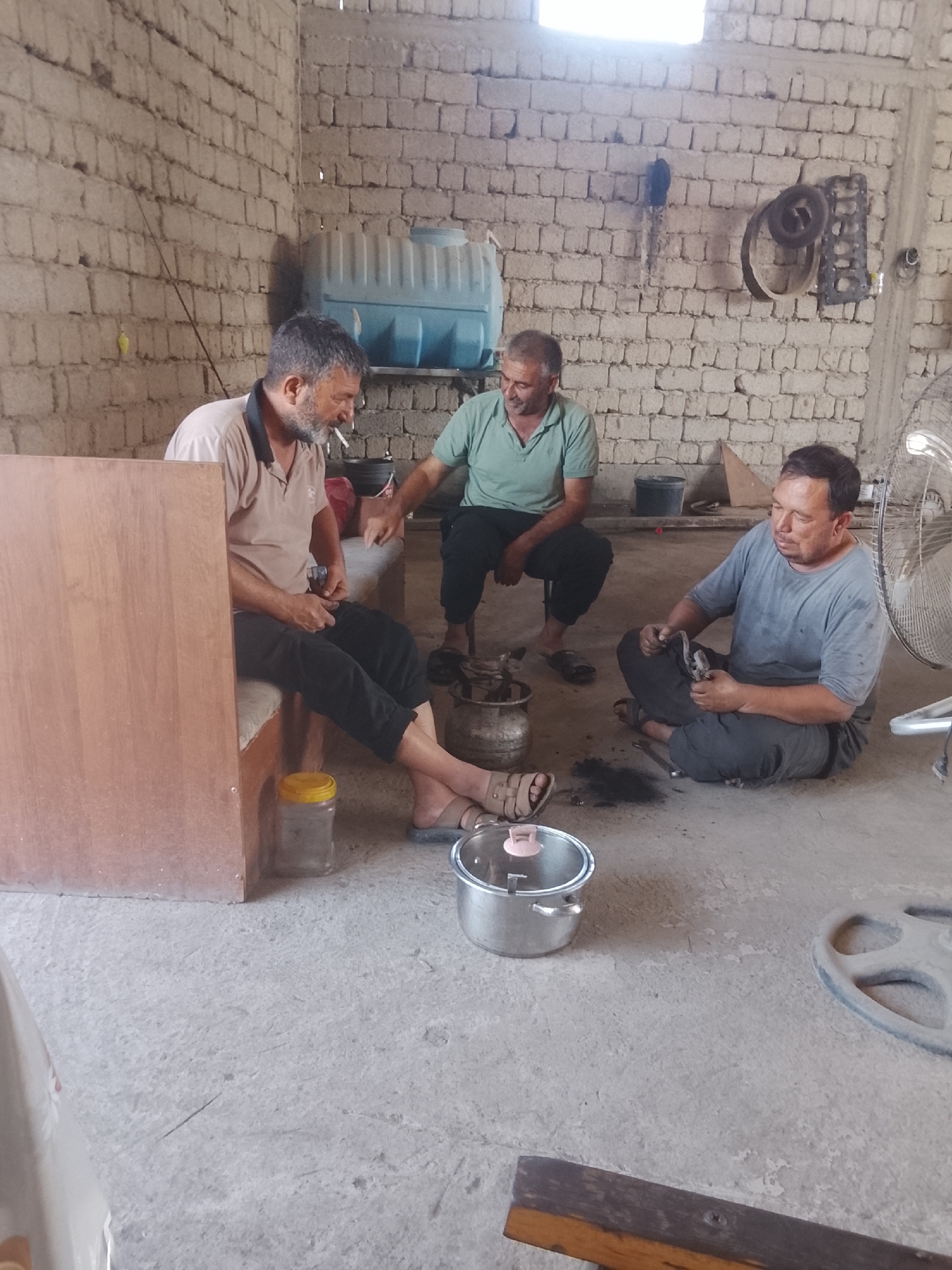 Three adult males are gathered inside a roughly constructed building with brick walls and a concrete floor. One man, wearing a beige polo shirt and dark pants, is seated on a bench and smoking a cigarette. He is holding a small metal object in his hands. To his right, another man in a green polo shirt and dark pants is seated on a stool, gesturing with his hands. A third man, wearing a gray t-shirt and dark pants, is sitting on the floor, holding a tool and appearing to work on something. Various objects are present in the environment, including a blue water tank on a shelf, a fan, a metal pot on the floor, and what appears to be scrap metal and tools scattered around. The overall impression is of a workshop or storage area in a functional, unadorned setting. The location is identified as Sabir Rakhimov, Uzbekistan.