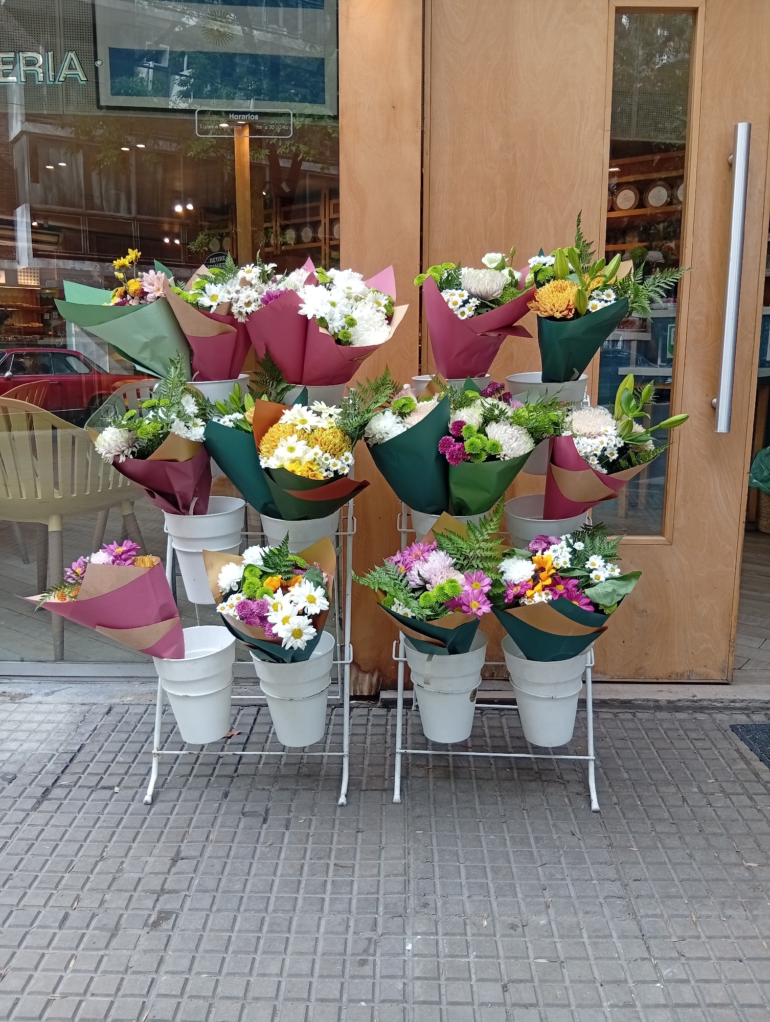The image captures a flower display outside a business in Buenos Aires, Argentina. The flowers are arranged in bouquets and placed in white buckets, which are set on a tiered, white metal stand. The bouquets are wrapped in green and maroon paper. The backdrop consists of a storefront window with a reflection of a red car and interior details, and a wooden door. The sidewalk is paved with gray square tiles. The name of the store "ERIA" is visible on the storefront. A small window sign is visible on the front glass. The display is well-lit, suggesting daylight. No people are visible in the immediate frame.