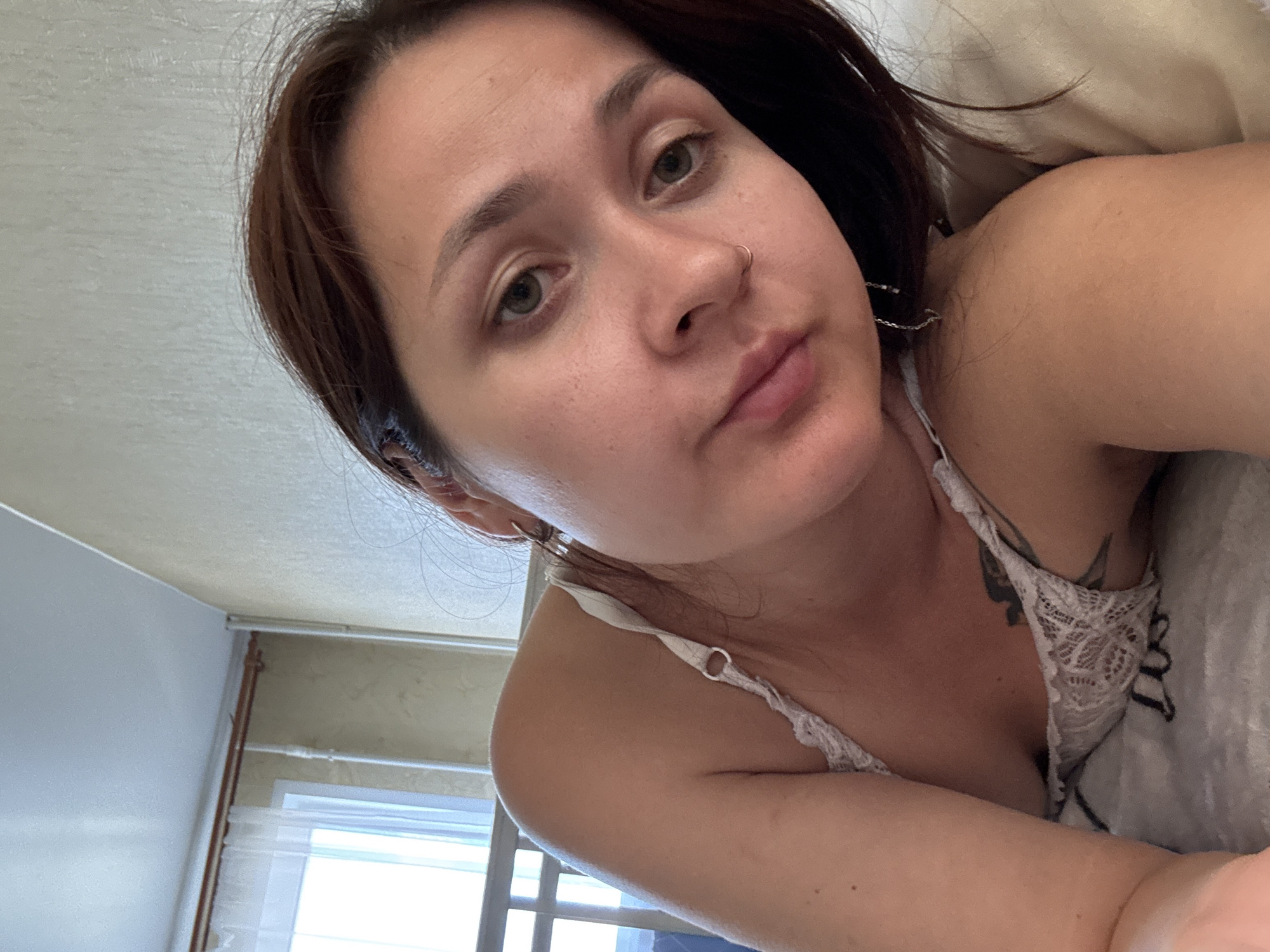 A young woman with brown hair and light skin is lying on a bed, looking at the camera. She is wearing a white lace bralette. A tattoo is visible on her chest. The room appears to be a bedroom with a window in the background and a white headboard. The lighting suggests it is daytime. The image does not provide specific details that would allow for the determination of its location within the city of Naberezhnye Chelny, Russia, or any other specific city.
