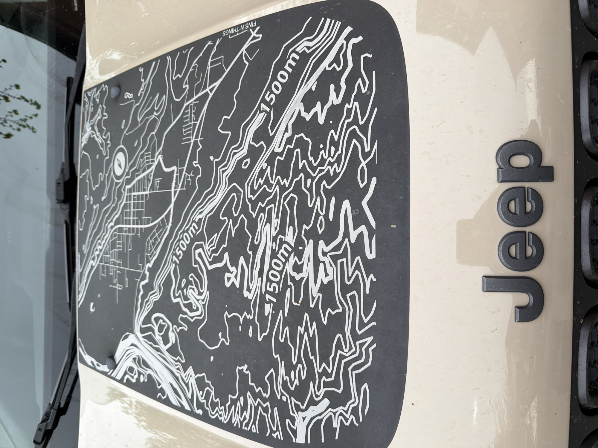 The image depicts a section of a beige Jeep hood, featuring a matte black topographic map sticker. The map includes contour lines, marked with "1500m," and what appears to be a compass rose. The sticker also displays the text "FINS N THINGS." To the right of the sticker, the black "Jeep" logo is attached to the hood. The windshield and a wiper blade are visible on the left. The map appears to represent an area with varying elevations and possibly a settlement or town, potentially indicating Dusheti, Georgia.