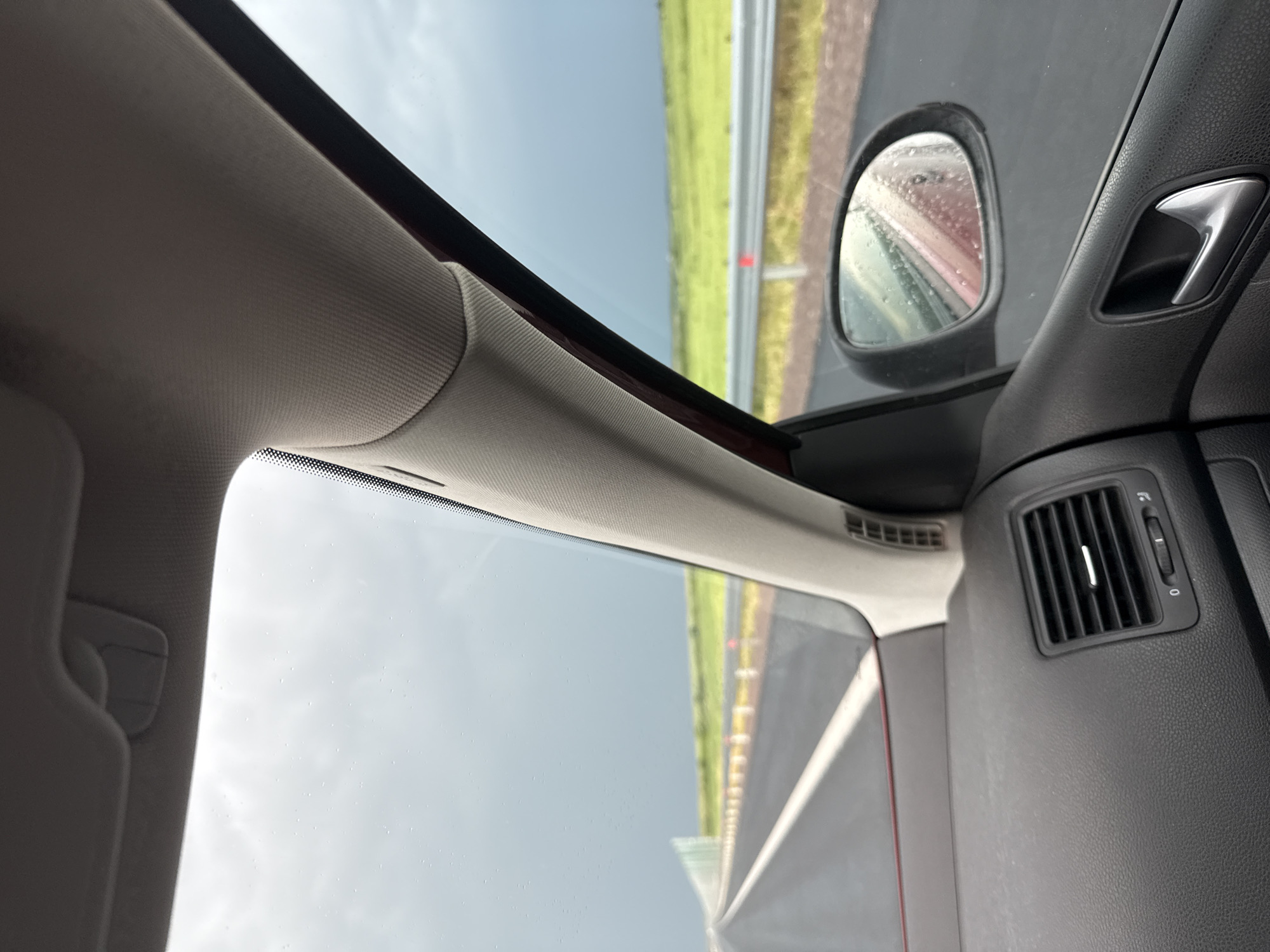 An eye-level interior shot from a vehicle captures a dynamic view, rotated ninety degrees clockwise, from the passenger side in Chokoba, Bulgaria. The car's A-pillar and part of the headliner, upholstered in light beige fabric, frame the left side and top of the image. A light-colored sun visor hangs partially into view on the far left.
Through the car's windshield and passenger window, an overcast sky is visible, with faint water droplets adhering to the glass, suggesting recent or ongoing rain. Beyond the window, a green, grassy expanse runs alongside a grey road or highway, which appears vertically oriented due to the image's rotation. Parts of a barrier or fence are also discernible along the road.
The right side of the image reveals elements of the car's dark grey dashboard and door panel. A textured black air vent with a vertical slider, faintly marked "0" and "1," is visible on the dashboard. Above it, a metallic door handle is integrated into the door panel. The side mirror, covered in numerous water droplets, shows a blurred reflection of the wet environment and a reddish hue, likely from the car's body. The overall scene depicts a journey in wet conditions, viewed from inside the moving or parked vehicle. No people are visible.