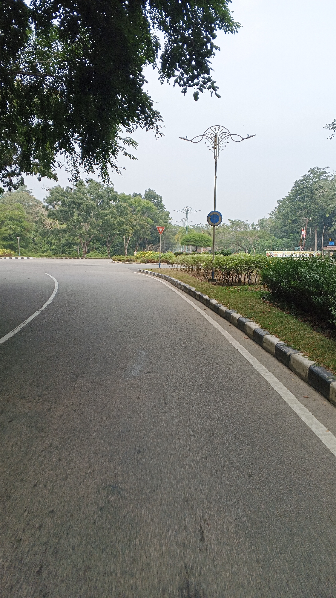 The image shows an asphalt road with a white line marking the lane and a black and white checkered curb. The road curves to the left, with a median strip on the right side of the road. The median strip is covered with grass and low-lying bushes. There are several trees and foliage in the background, along with a lamppost and traffic signs. The sky is overcast. The scene appears to be located in a park-like setting within the city of Kampung Rempak, Indonesia.
