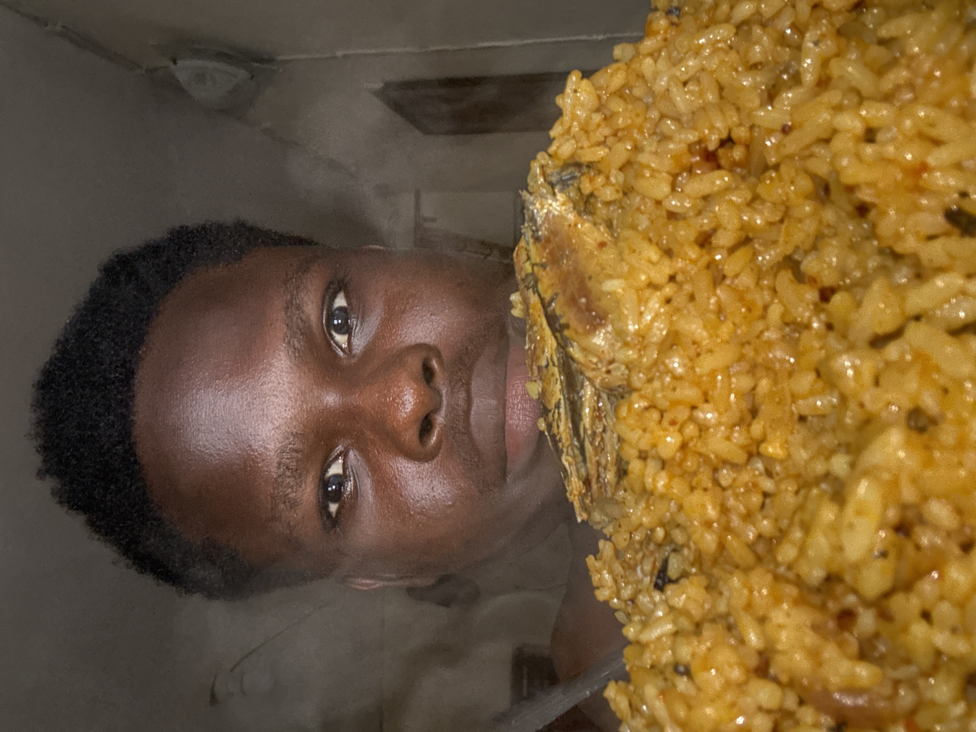 A young man with short, dark, curly hair and dark skin is partially visible, positioned behind and looking over a large quantity of food. His forehead, eyes, nose, and upper lip are visible. His skin appears slightly oily, reflecting light. His dark eyes are directed forward. A metallic utensil, possibly a spoon, is partially visible on the far left. The lower half of the frame is dominated by a mound of reddish-orange rice, consistent with jollof rice, from which steam is faintly rising. Several pieces of cooked fish, with dark and golden-brown skin, are placed on top of the rice. The background is dark and indistinct, suggesting an indoor setting. On the right, a light-colored fixture is visible on a textured wall, and a darker rectangular element, possibly a doorframe, is also present. The scene is located in Ihiagwa, Nigeria.