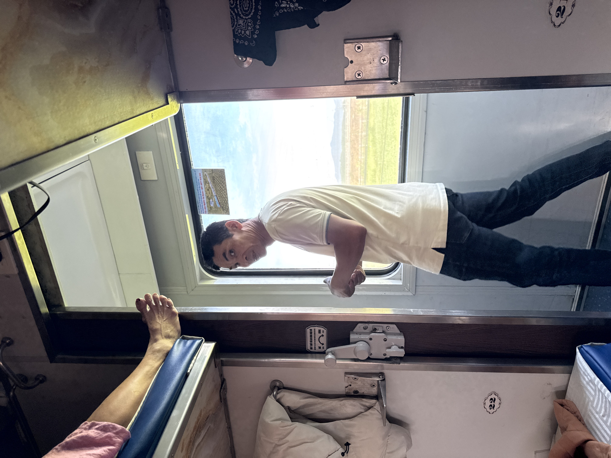 The image shows the interior of a train compartment with a man in a light-colored polo shirt and dark jeans walking towards the camera. He is looking back over his shoulder. To the left of the man, a person's bare foot is visible resting on a blue bunk bed. The compartment has multiple bunks, with blankets and bags stored on them. A window to the right reveals a bright, sunny outdoor scene with a green field and distant mountains. The train appears to be in motion, as suggested by the landscape outside. The overall impression is of a functional and somewhat utilitarian train travel experience. The location is identified as Bình Nguyên, Vietnam.