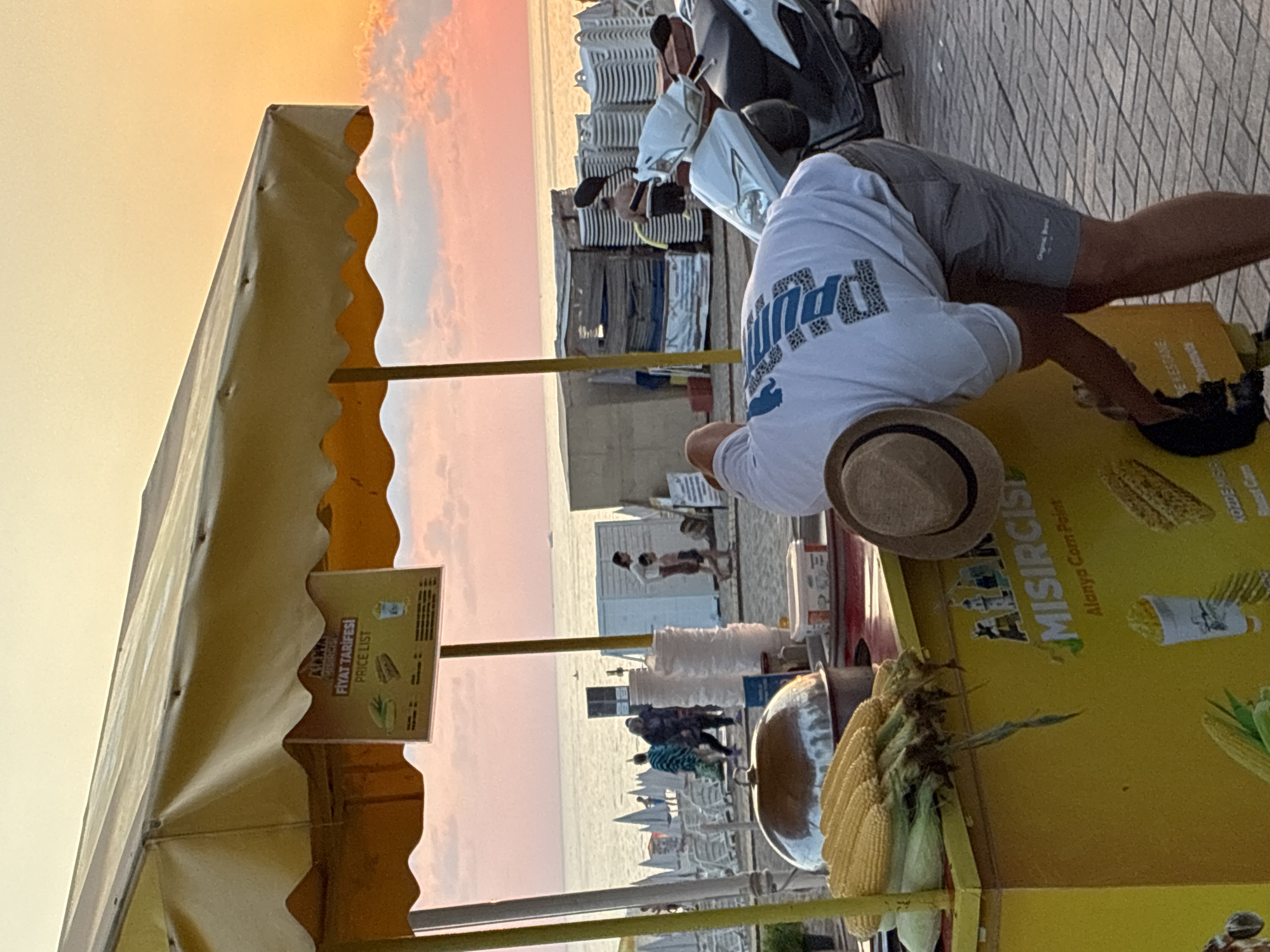The image depicts a beachside corn stand in Alanya, Turkey, during sunset. A person, wearing a white t-shirt and grey shorts, is bent over the yellow corn stand, appearing to be preparing or serving corn. The stand has a yellow awning and displays images of corn and other food items. In the background, the sea is visible with several lounge chairs set up on the beach, and a few people are walking along the shore. Two scooters are parked near the stand, and the overall atmosphere suggests a casual evening on the coast. The sky is a warm blend of orange and pink hues, characteristic of a sunset.