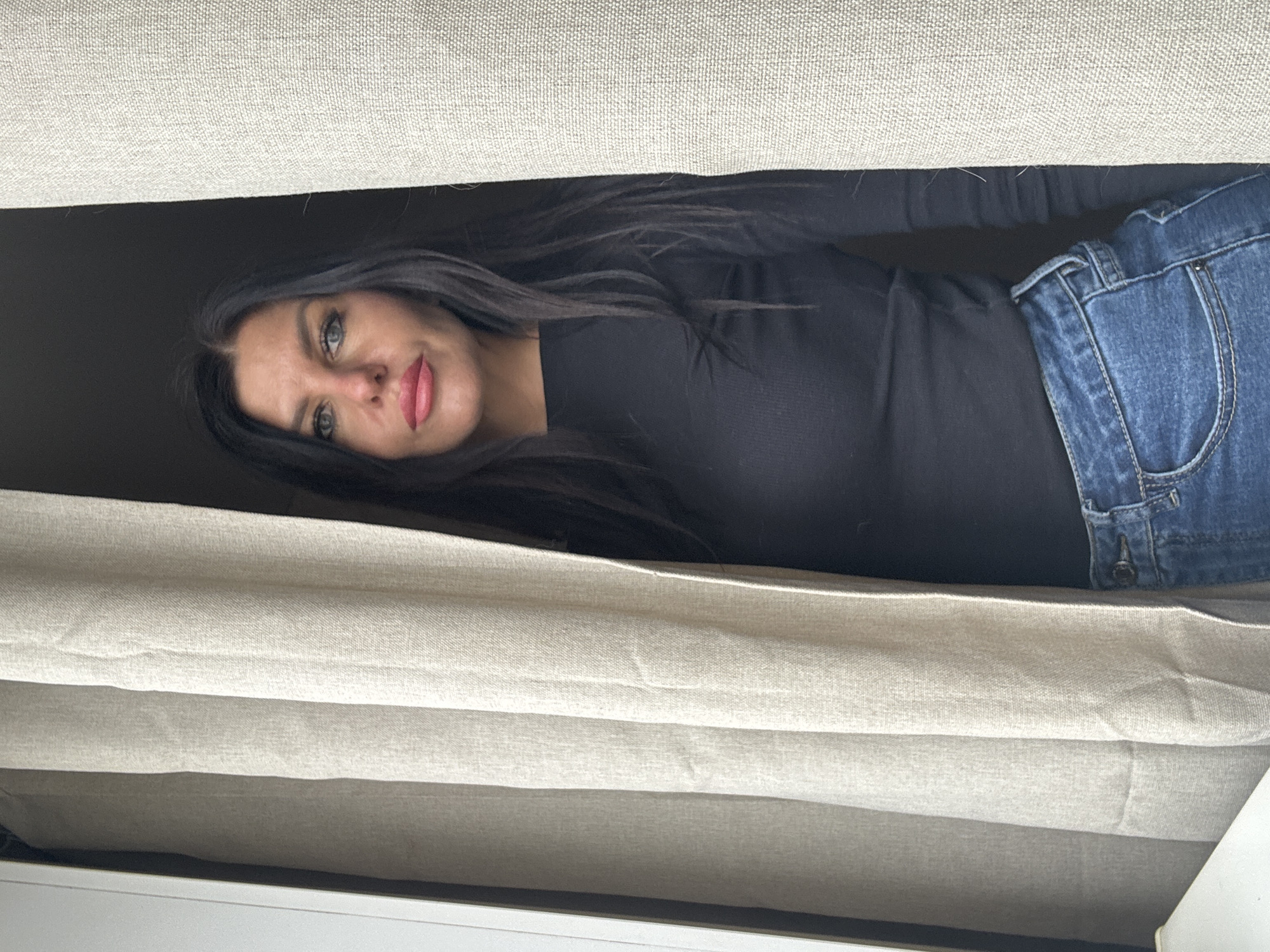 A person, likely a woman, is peering through an opening in window blinds. She is wearing a black long-sleeved shirt and blue jeans. She has dark hair and fair skin. The background behind her is dark, potentially indicating an interior space. The location is not discernible from the image.