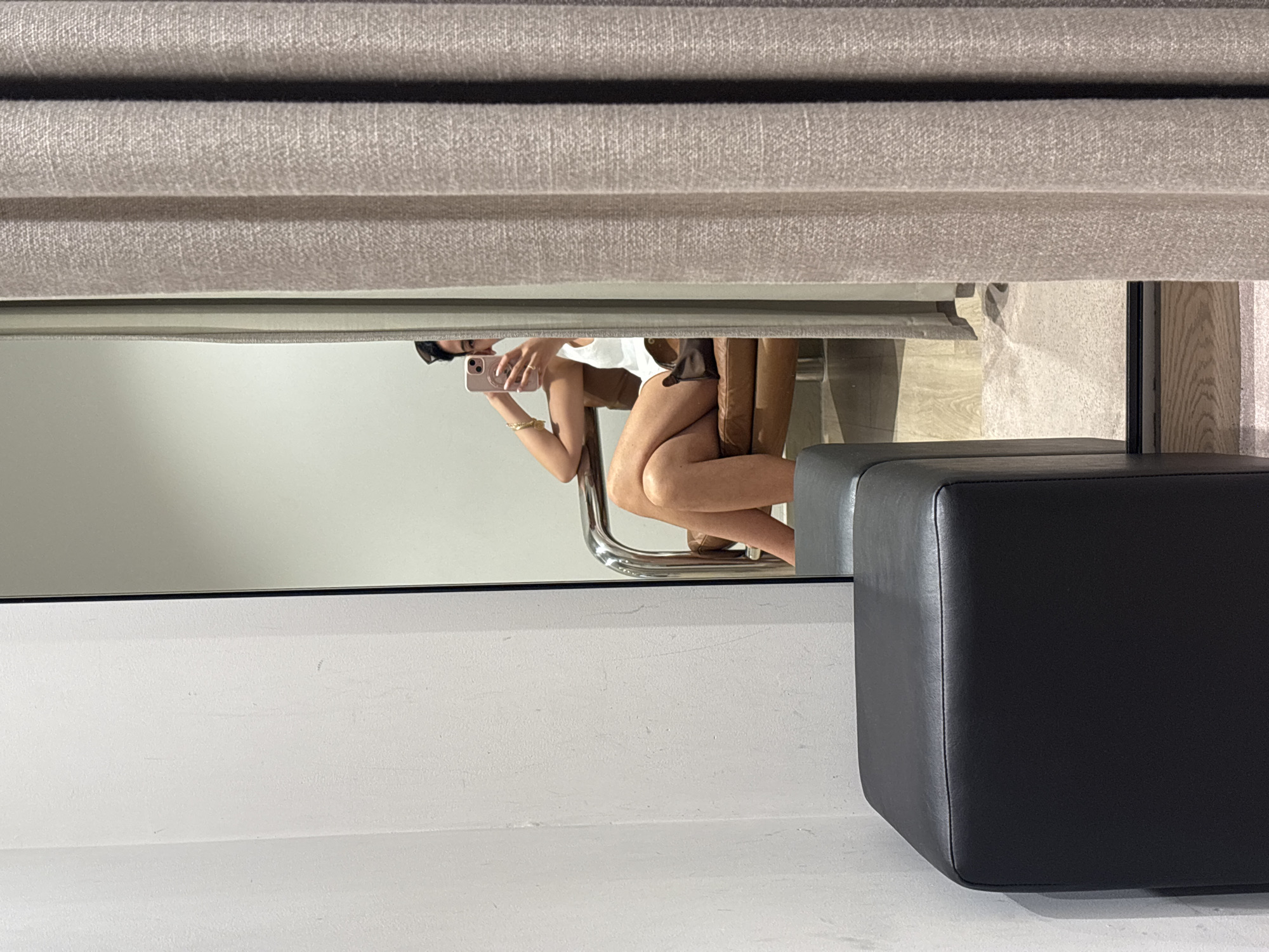 The image presents a reflection in a mirror, likely within a clothing store fitting room. A person is visible in the mirror, seated in a chair and holding a mobile phone. The person appears to be taking a photograph of themselves. The fitting room contains a black, square ottoman, visible in the foreground. The walls of the fitting room are a neutral, light color. A set of light brown curtains is visible at the top of the image, partially obscuring the reflection. The specific location in London, United Kingdom, cannot be determined from the information present in the image.