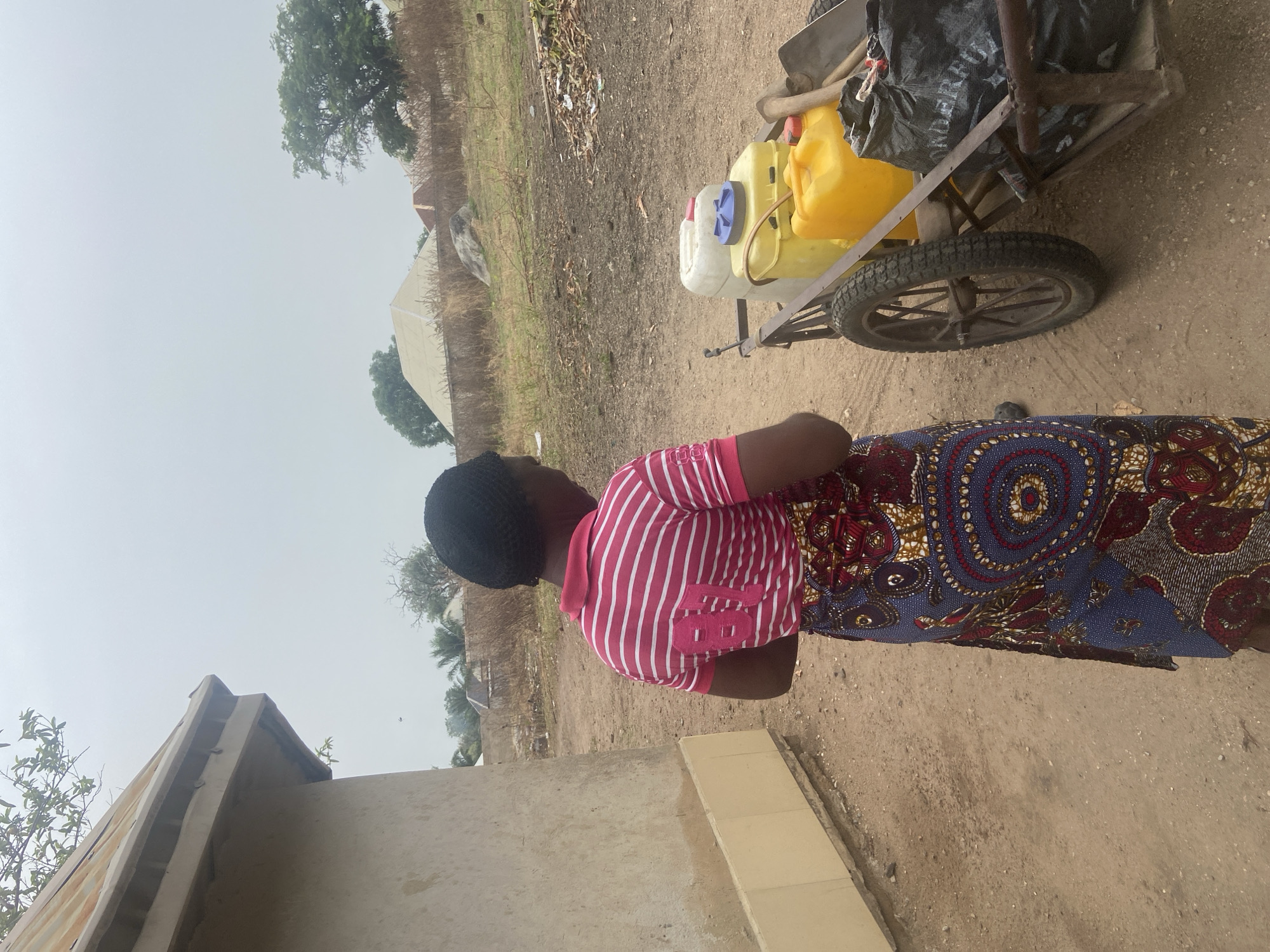 This image, taken in Mararaba, Nigeria, shows a woman standing next to a two-wheeled handcart on a dirt path. The woman is viewed from her back-left side, facing away from the camera. She wears a black head covering, a red and white striped short-sleeved top with the number "78" (or "7B") printed in white on the back, and a long, colorful patterned skirt or wrap, likely made of Ankara fabric, featuring blue, red, and golden patterns.
The handcart, positioned to her right, is laden with several items. Most prominent are two large yellow plastic jerrycans and one white jerrycan, typically used for carrying water. A black plastic bag is tied to the cart, partially obscuring other contents, and what appears to be the handle of a shovel or similar tool is also visible, propped against the cart's frame. The cart has sturdy, spoked wheels, suitable for unpaved terrain.
The setting is a rural or semi-urban environment. The ground is unpaved dirt, and sparse vegetation, including trees, can be seen in the background. In the distance, several simple structures or huts are visible. The sky is overcast or hazy, indicating diffused light with no strong shadows, suggesting either an early morning, late afternoon, or a generally cloudy day. The scene captures an everyday activity likely related to water collection or transport.