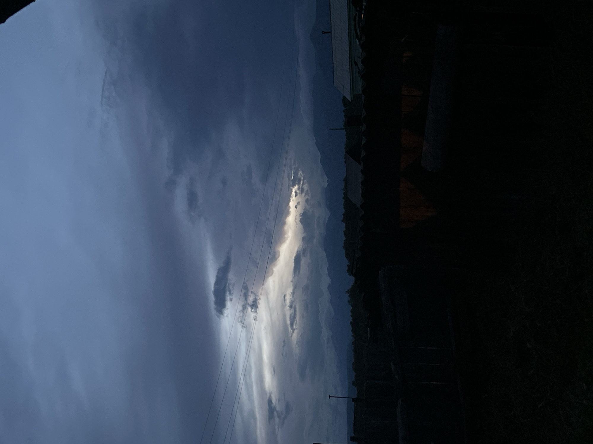 The image depicts a dark, overcast sky with dramatic clouds, hinting at twilight or a storm. A sliver of bright light pierces through a gap in the clouds, illuminating the distant, hazy silhouette of a mountain range. In the foreground, the tops of several dark, indistinct structures are visible, possibly rooftops or fences, suggesting a rural or suburban setting. Power lines stretch horizontally across the upper portion of the frame. The overall mood is somber and atmospheric, with limited visibility due to the low light conditions.