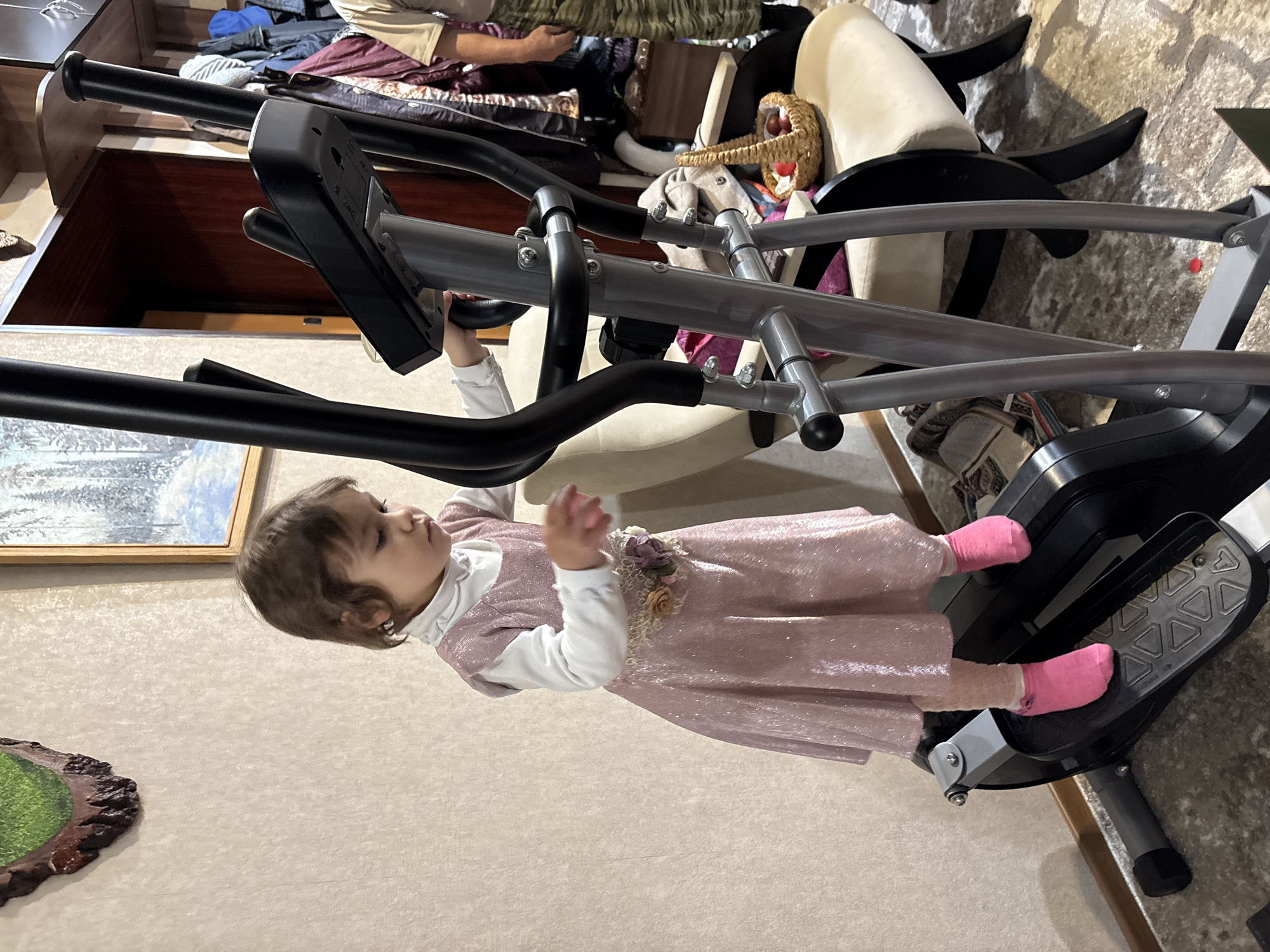 A toddler in a pink dress and pink socks stands on the pedal of an elliptical machine, holding onto the handlebar. The environment is cluttered, with furniture and miscellaneous items visible in the background. The location is impossible to determine as there are no locational visual clues.