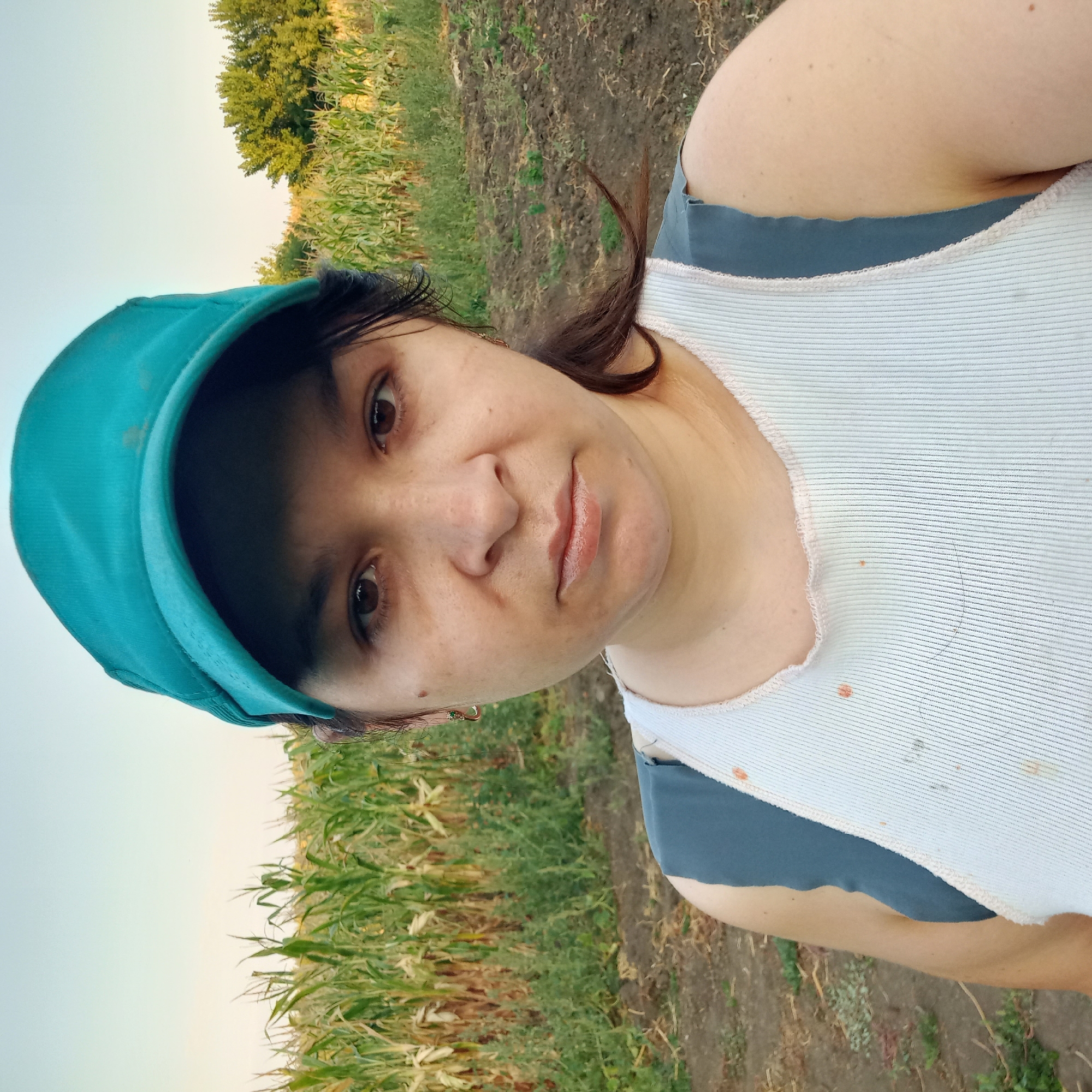 In the city of Klyntsi, Ukraine, a woman in a teal baseball cap and a white tank top stands in front of a cornfield. The woman's face is directed towards the camera. The background shows a field of corn and trees under a clear sky. The ground in the foreground is dirt. The woman is wearing earrings and her hair is pulled back. The tank top has some stains on it.