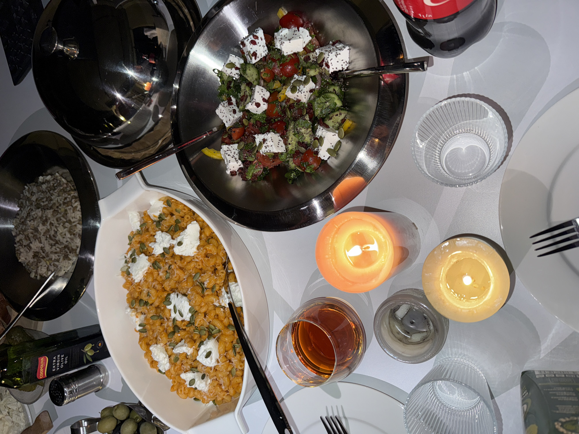 The image depicts a dinner table setting, likely in a private residence, with a variety of dishes and items arranged on a white surface. There is a large silver bowl filled with a salad composed of tomatoes, cucumbers, peppers, and feta cheese, all dressed with seeds. Another white bowl contains pasta with pumpkin seeds and cream cheese. Nearby is a smaller bowl with rice.
A bottle of Coca-Cola is visible, along with a wine glass filled with a light-colored liquid. There are two lit candles, one with an orange hue and one with a yellow hue, along with an empty candle container.
Other items include a bottle of olive oil, a salt shaker, a dish of olives, and place settings consisting of plates, silverware, and glassware.
The location is not discernible from the image. Thus, determining if the picture was taken in Yerevan, Armenia, is impossible based solely on the visual information. There is no evidence of people, interactions, or specific actions happening.