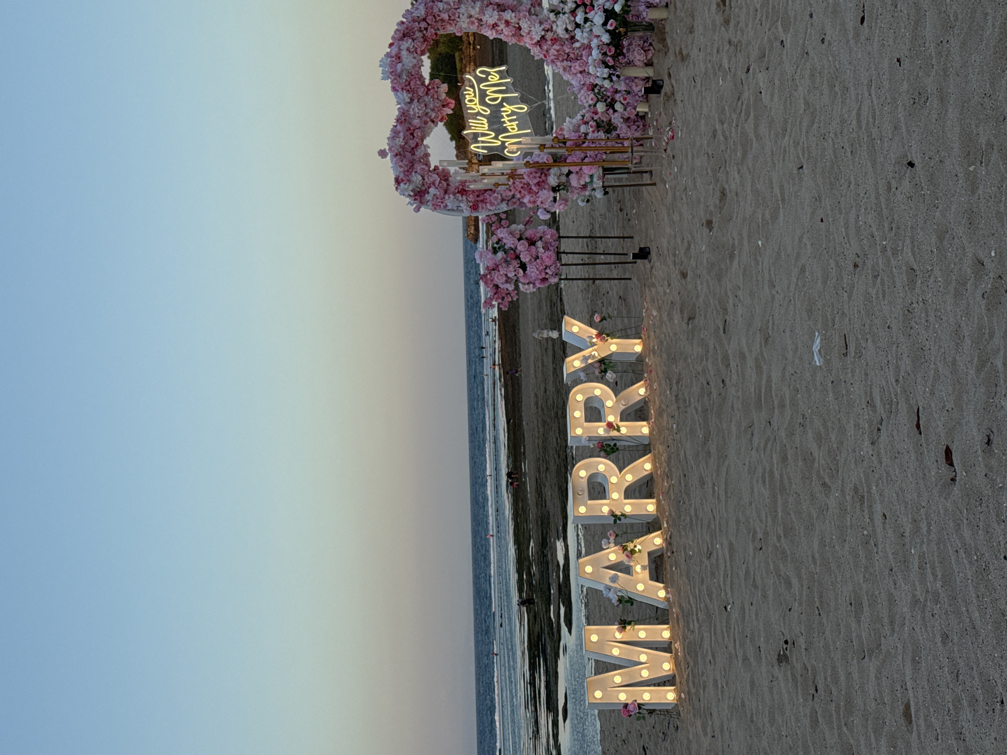 This image depicts a romantic beach proposal setup on the coast of Kuta Selatan, Indonesia, likely during sunset given the warm light. In the foreground, large illuminated marquee letters spell out "MARRY" in capital letters, adorned with pink and white flowers. To the right, a large heart-shaped arch, also decorated with pink and white flowers, frames a neon sign that reads "Will you Marry Me?" in cursive script. Several tall, slender candles in clear holders stand within the heart arch, adding to the ambiance. Behind the proposal setup, the ocean stretches out, with gentle waves lapping at the shore. The beach itself is sandy, with scattered natural debris. In the mid-ground and background, several individuals are visible, some walking along the water's edge and others interacting near the shore, suggesting the presence of other beachgoers. The sky transitions from a soft blue near the horizon to a lighter hue towards the top of the frame.