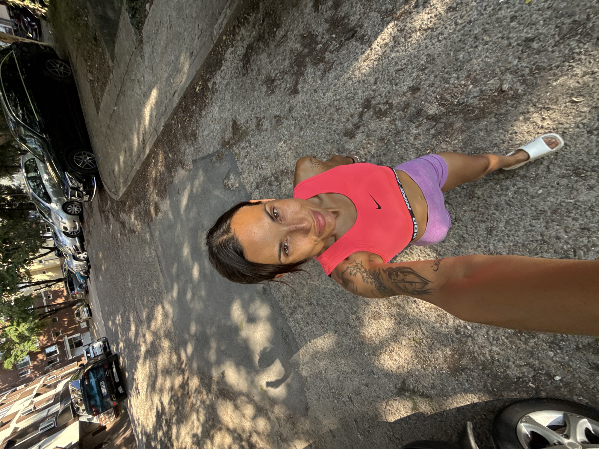 In the city of Pleven, Bulgaria, a woman is taking a selfie in an outdoor setting. The woman is wearing a bright pink sports bra and purple shorts. She has tattoos on her left arm and shoulder. She is standing on a gravel path. In the background, there are several parked cars, including a black Mercedes-Benz station wagon, a silver hatchback, and a dark blue SUV. A multi-story brick building is visible on the left side of the image. Trees are casting shadows on the ground, indicating it is a sunny day. The image is taken from a slightly high angle, looking down at the woman.