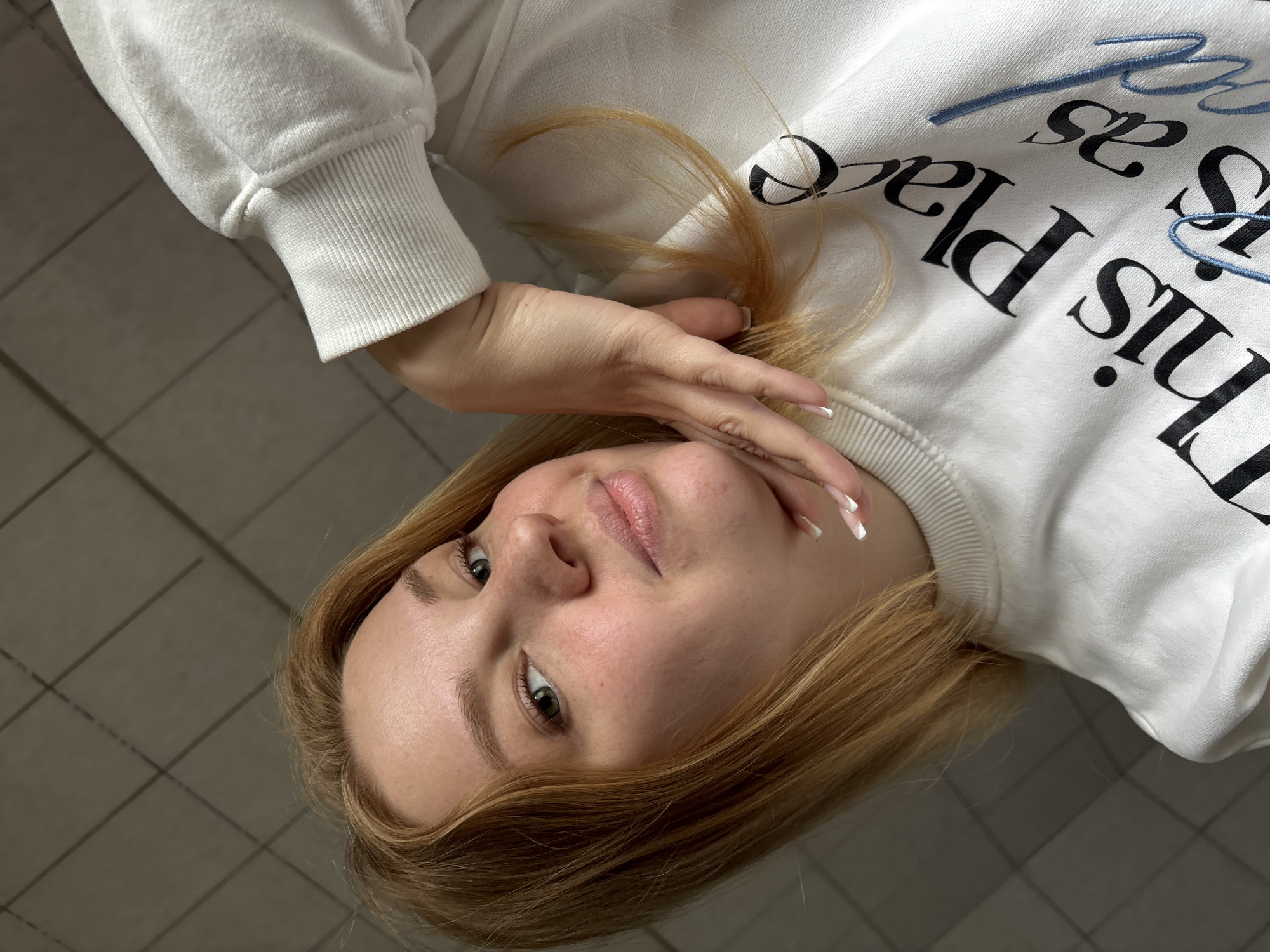 A young woman with blonde hair is lying down, looking up at the camera. She is wearing a white sweatshirt with black lettering. Her right hand is near her face, with long fingernails. The background appears to be a tiled floor. The location cannot be identified as Rusanovo, Russia, based on the image alone.
