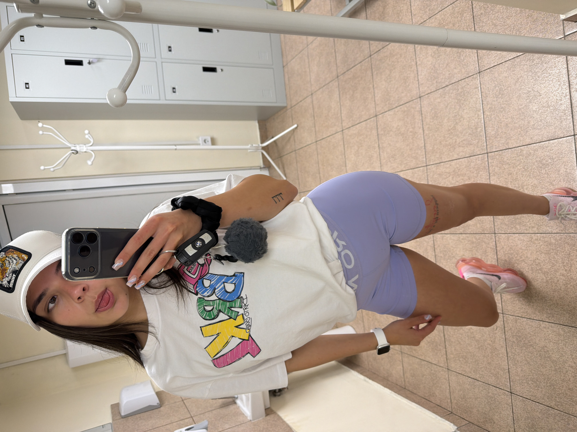 A young female, positioned in a mirror selfie, is observed in what appears to be a changing room or public facility within the city of Gabrovo, Bulgaria. She has dark hair, partially concealed by a white baseball cap adorned with a brown and white tiger patch on the front. Her head is tilted, and she is looking to her right while sticking her tongue out. She wears a white short-sleeved t-shirt featuring a stylized, multi-colored graphic that reads "YRKS" in pink, yellow, green, and blue, with smaller black text "GOOD TRACK" underneath. Her lower body is clad in light lavender-colored compression shorts with a visible waistband text "ROY" or "ROK" upside down. On her right wrist is a white smartwatch, and on her right upper arm, a small tattoo of three vertical lines is visible. Another tattoo is partially visible on her right thigh. She wears pink and orange athletic shoes. Her left hand holds a dark grey smartphone (likely an iPhone Pro model with triple lenses) that captures the reflection, along with a BMW car key fob featuring a grey fluffy pom-pom, and a black hair scrunchie. The background on the left displays part of a tiled wall with light brown/beige square tiles, a white sink, and a wall-mounted hand dryer. To the right, against a light beige wall, stands a white, multi-hook coat rack, near which a white power outlet is mounted. Further right, a stack of six white metal lockers with black handles and keyholes is visible. The floor throughout the scene is covered in light brown/beige square tiles with a speckled texture. The entire scene is brightly illuminated, suggesting artificial lighting.