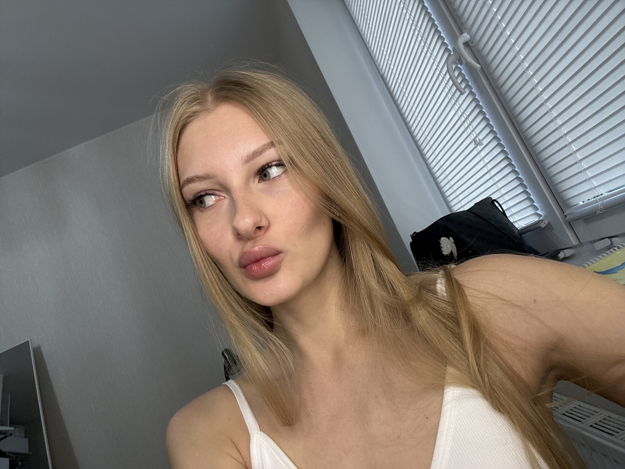 A young woman with blonde hair is taking a selfie indoors. She is wearing a white tank top. She is looking to her left with her lips puckered. Behind her, there is a window with blinds and a black bag. The walls are painted a light gray. The location is not specified in the image.