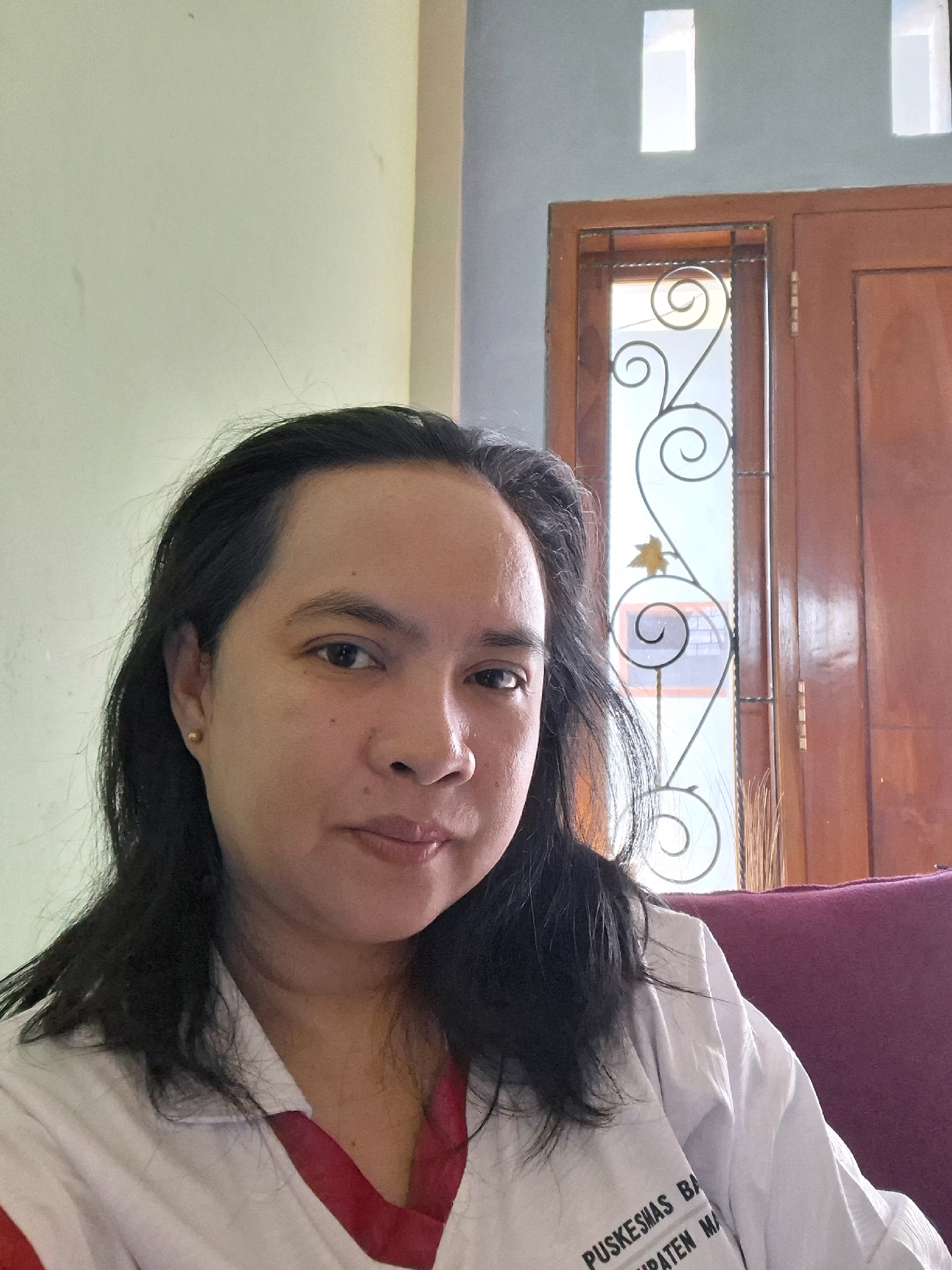 In the city of Bantur, Indonesia, a woman with long, dark hair and a fair complexion is pictured. She is wearing a white shirt with red trim and some text visible on it. The text appears to be "PUSKESMAS BA" and "MPATEN MA", possibly indicating she is affiliated with a community health center. She has a small gold stud earring in her left ear. The background shows a light-colored wall, a wooden door with decorative ironwork on a glass panel, and two small rectangular openings near the ceiling that let in light. A purple upholstered surface, likely a couch or chair, is partially visible on the right side of the image. The lighting suggests it is daytime.