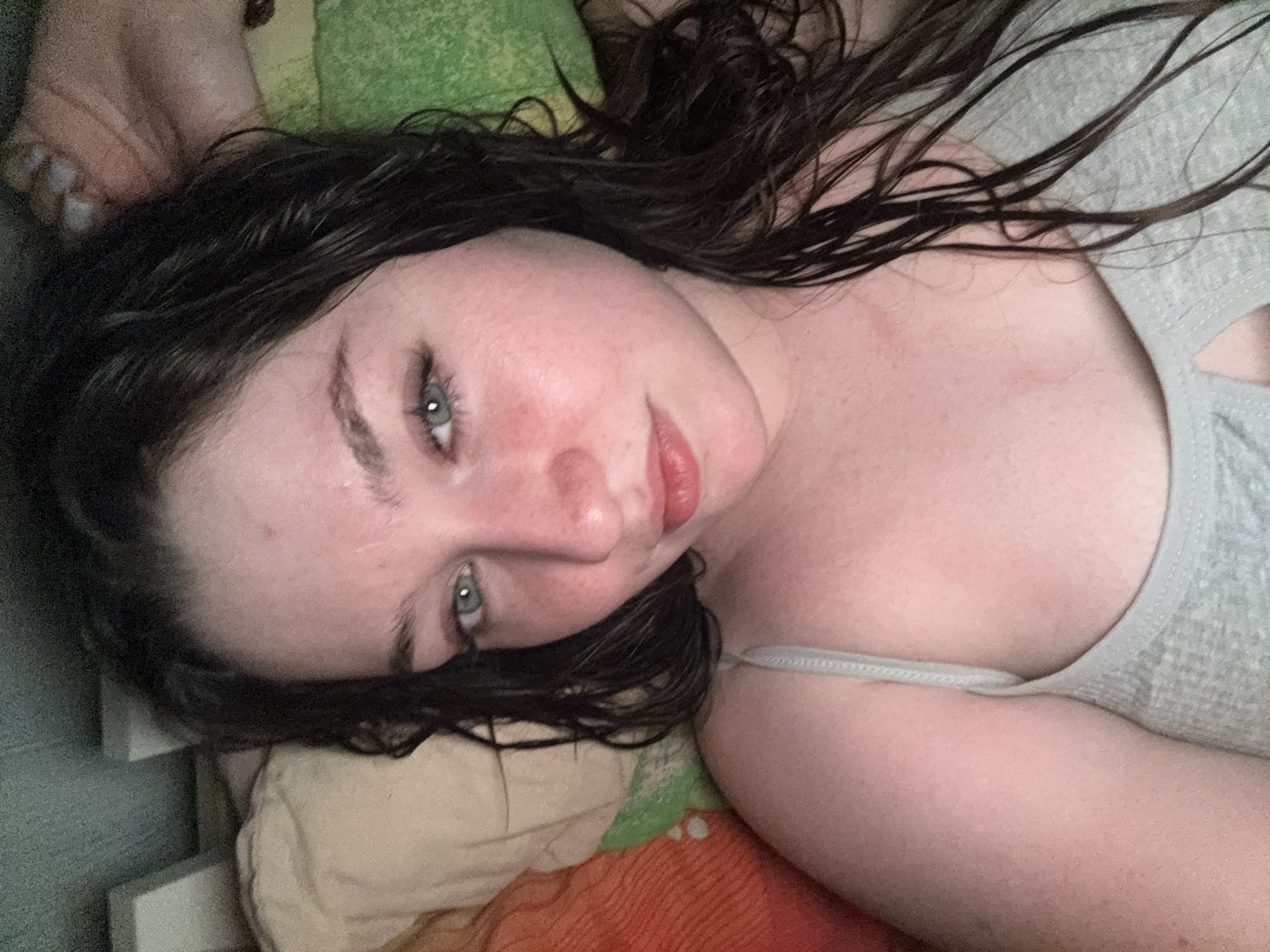 A young woman with dark hair and light green eyes lies in bed, looking at the camera. She is wearing a grey camisole. Her face is slightly flushed. The background shows pillows and bedding. The setting is a bedroom in Бородулино, Russia.