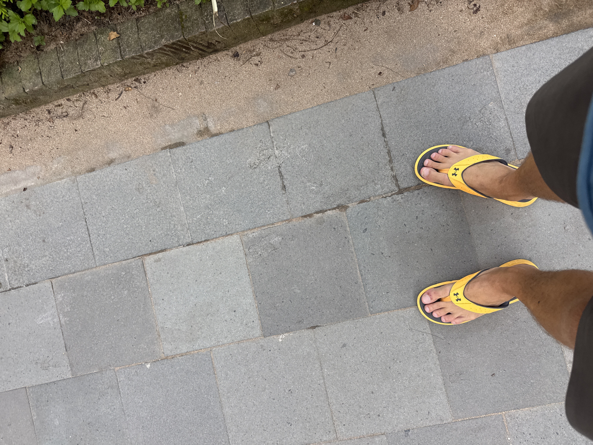 The image shows a first-person perspective of a person standing on a gray-tiled walkway. The person is wearing yellow and black Under Armour flip-flops. Part of the person's legs, covered by gray shorts, is visible on the right side of the image. To the left of the walkway is a border of tan-colored sand or dirt, followed by a raised brick edge and some greenery. The location is Denpasar, Indonesia.