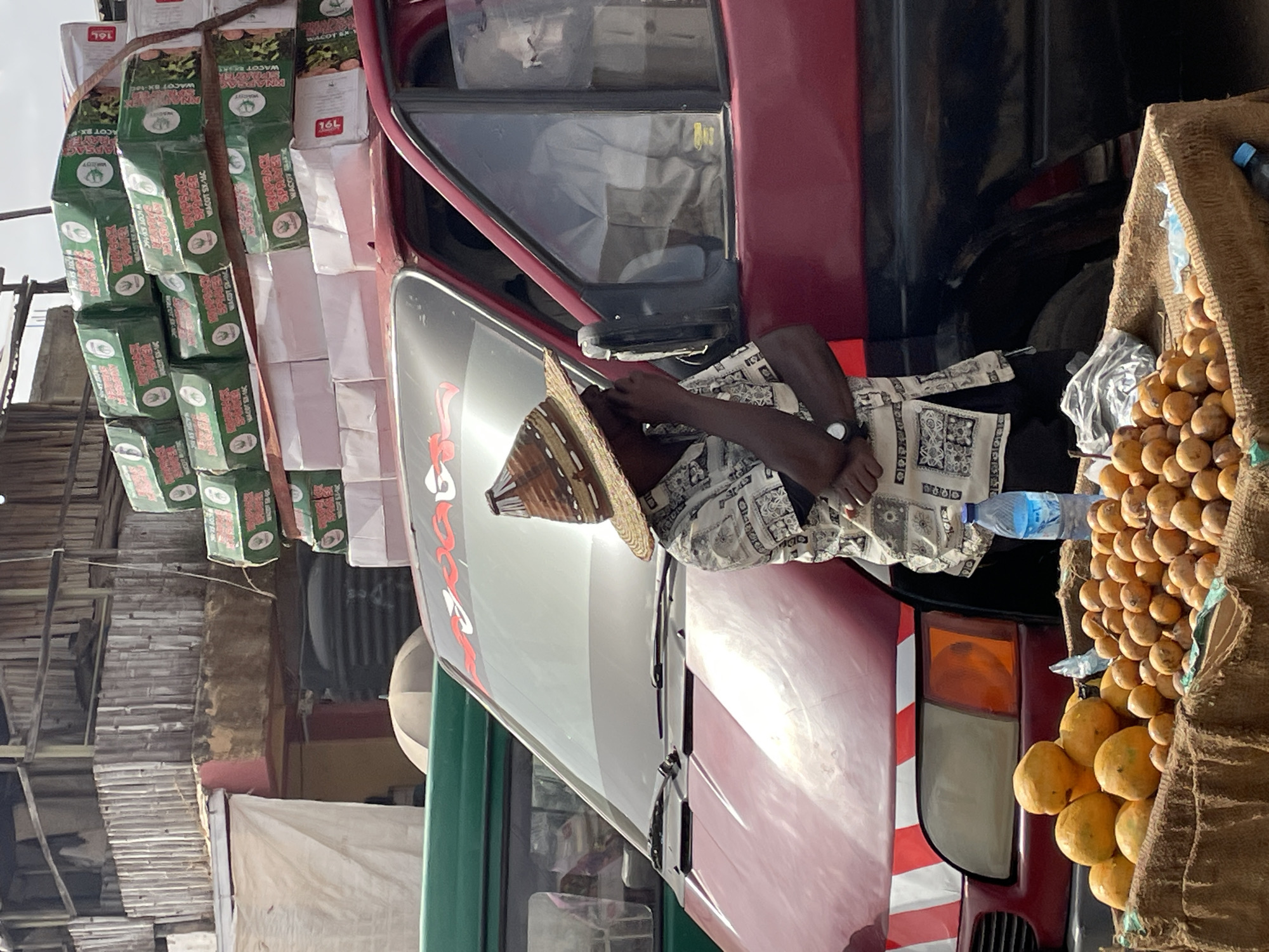 The image captures an outdoor scene, likely a market or roadside, centered around a loaded red and black vehicle. A dark-skinned person, wearing a wide-brimmed conical straw hat and a patterned shirt, is leaning against the side of the vehicle near the driver's window, with a wristwatch visible on their left arm. A clear plastic water bottle is placed beside them.
The vehicle, possibly a truck or large van, is heavily laden with goods. Numerous green and white boxes are stacked high on its roof and along its side. Visible text on these boxes includes "WACOT," "WACOT SX-14C," "KNAYBAK," and "16L," suggesting commercial products or agricultural supplies.
Adjacent to the person, a large woven basket overflows with many round, yellowish-orange fruits, which appear to be oranges or similar citrus. The background reveals rustic structures made of wood and what looks like corrugated metal, implying a bustling, possibly informal, commercial setting. The bright, natural light indicates it is daytime. The overall activity suggests the transportation and sale of goods.