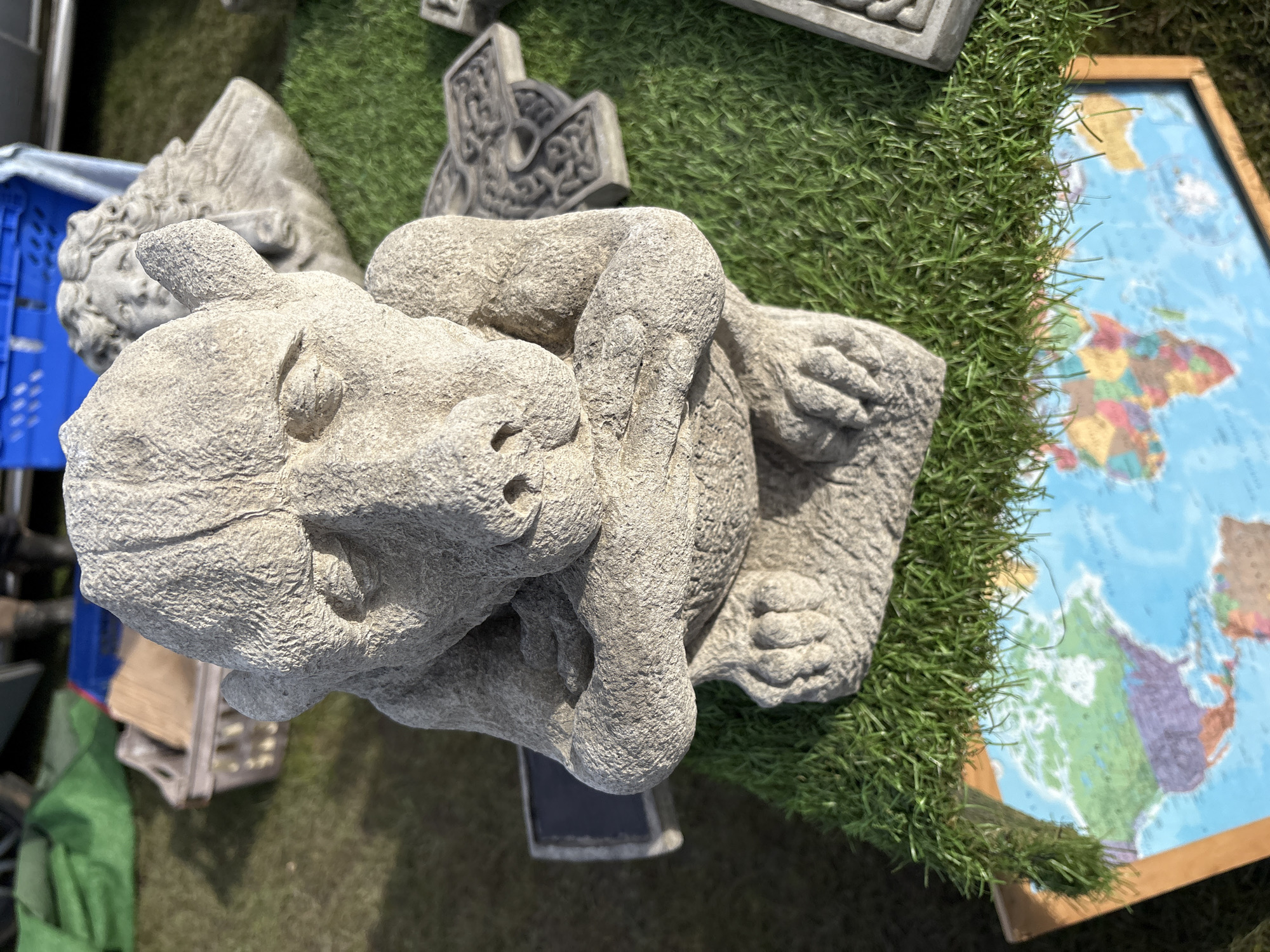 A high-angle, full-color photograph shows a collection of decorative items, potentially for sale, resting on the ground in Chirk, United Kingdom. A light gray, concrete-like gargoyle statue with folded arms is prominently positioned in the foreground, adjacent to a patch of artificial turf. To the right of the statue and turf is a framed world map with colorful continents on a blue ocean background. Other concrete decorations of varying shapes and sizes are scattered around, along with a blue plastic crate. The ground appears to be a grassy or dirt surface, suggesting an outdoor setting. The scene is captured in daylight, with the sun casting shadows on the objects.