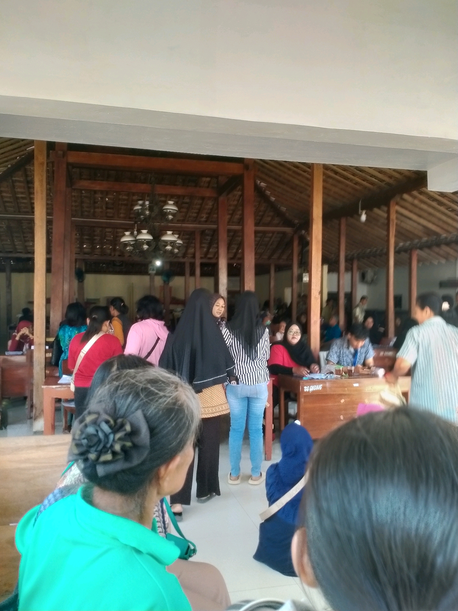 The image depicts an outdoor gathering at what appears to be a community event or meeting in Playen, Indonesia. A group of people, predominantly women, are seen interacting in a covered, open-air pavilion with traditional Indonesian architecture, characterized by wooden pillars and a thatched roof. The atmosphere seems relaxed, with individuals engaged in conversation and observation. The scene is well-lit, suggesting it is daytime. The foreground is dominated by the backs of two women, one in a vibrant green top and the other with dark hair. Further back, several other women are visible, some standing and others seated at wooden tables. One woman in the center is wearing a black hijab and a patterned skirt, while another to her left wears a pink top and a black strap across her chest. A person in a blue hijab is kneeling near the tables. In the background, men and women are seated at tables, possibly attending to administrative tasks or participating in the event. The presence of a "DS GADING" label on a table suggests a location within the Gading village area. The overall impression is of a local community engagement taking place in a rustic, open-air setting.