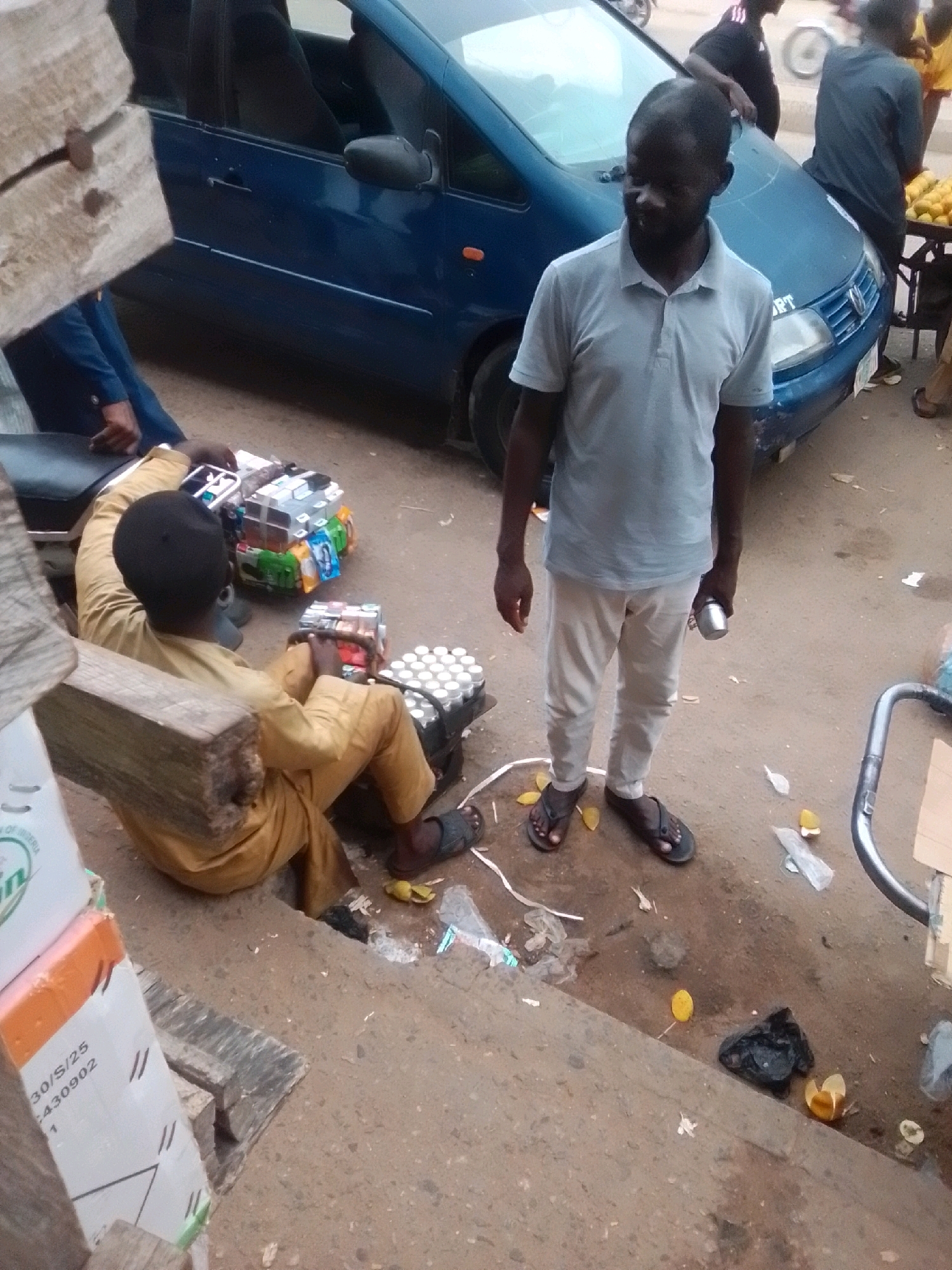 In Arawa, Nigeria, a man stands on a dusty street next to a blue van, holding a metal cup. He is wearing a light gray polo shirt and cream-colored pants. Another man, seated on a wooden bench, is dressed in a yellow outfit and appears to be arranging stacks of canned beverages. Several cases of drinks are visible around him. The scene depicts a commercial setting, possibly a roadside stall, with a general appearance of a typical urban environment in Nigeria.