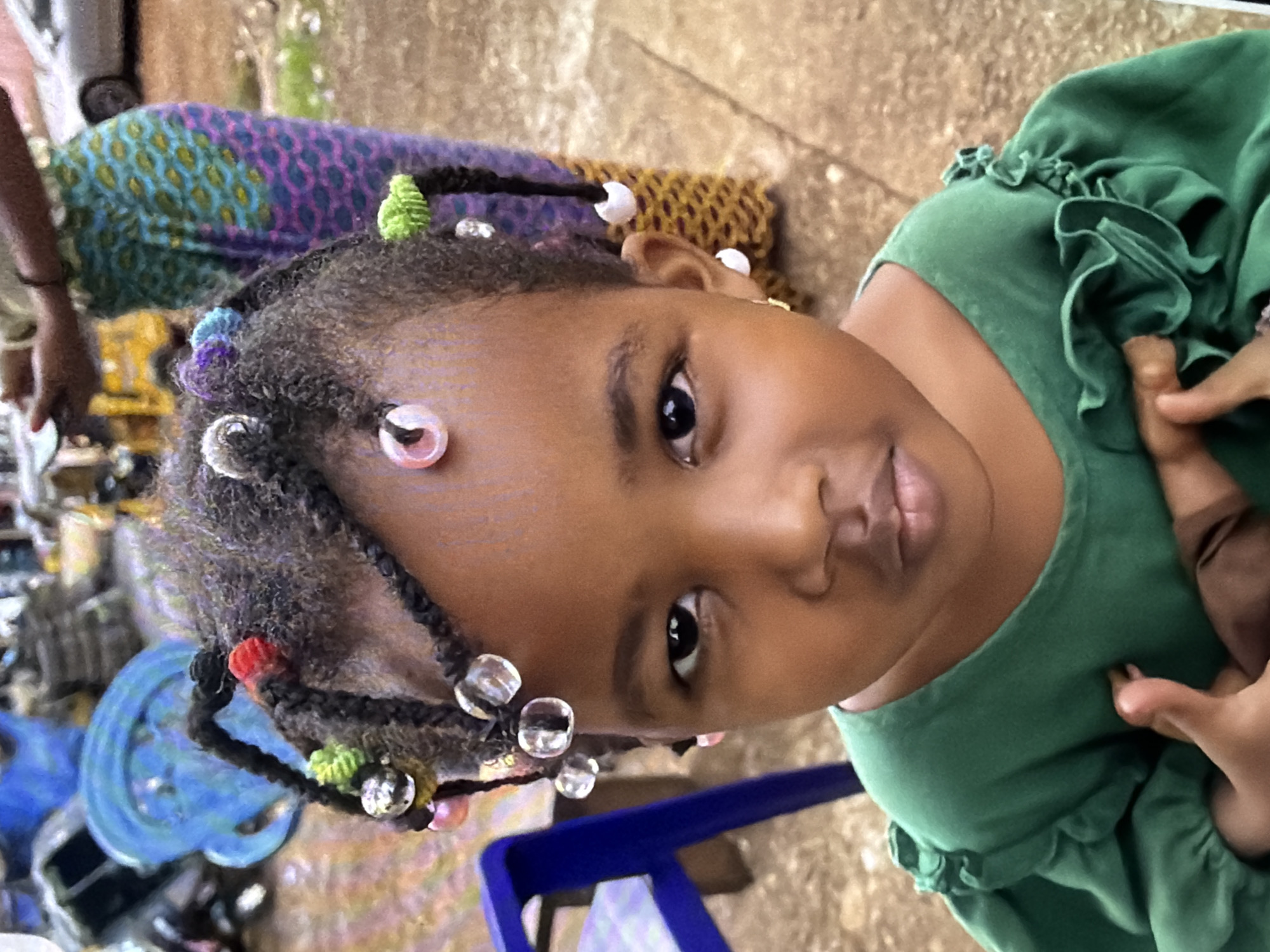 A close-up, high-angle shot captures a young Black girl from the chest up. She is wearing a green, ruffled-sleeve dress and has her hair styled in braids adorned with colorful beads and shiny accents. The background is a mix of earthy tones, with a blurry pattern of purple and green fabric visible behind her left shoulder. The girl's expression is neutral, and her eyes are focused slightly to her left. The lighting appears to be natural, suggesting an outdoor setting during daylight hours. The overall mood is casual and everyday, with no specific event or activity clearly depicted. The image is composed to highlight the girl's face and hair details.
