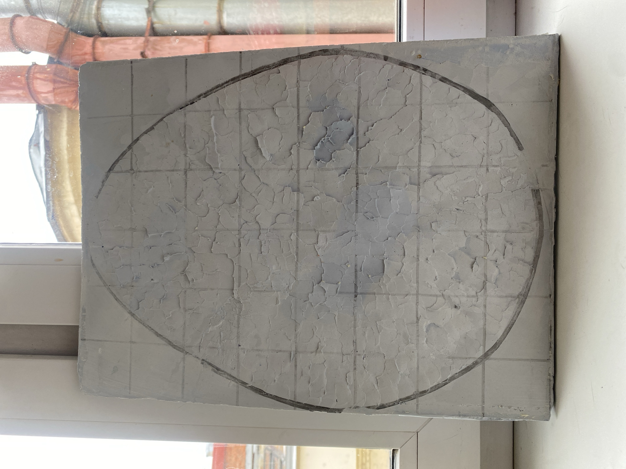 A square, gray, textured tile or panel rests on a white windowsill inside what appears to be an apartment. The tile has a grid pattern lightly inscribed on its surface and a roughly drawn oval shape in black outlining the central area. The texture of the tile's surface is cracked and uneven. Through the window, elements of the outside environment are visible, including pipes, a building in the distance, and a view of the sky. The precise location in Moscow, Russia cannot be determined from the image alone.