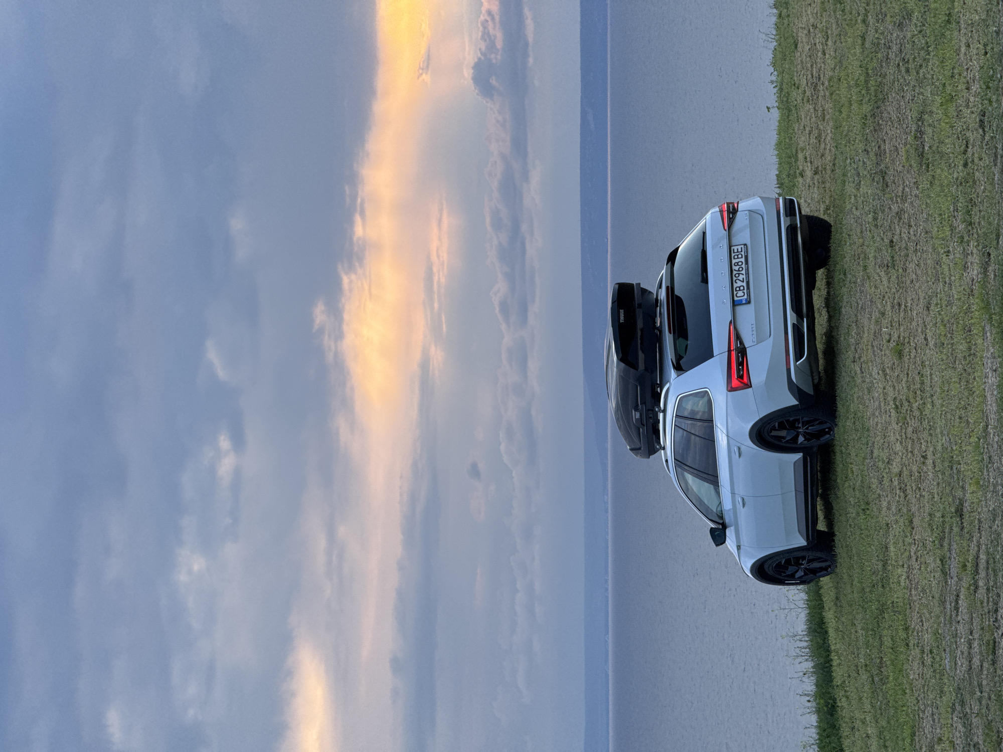 The image shows a white Skoda Karoq parked on a grassy field in what appears to be Balgarevo, Bulgaria. The car has a Thule roof box. The license plate reads "CB 2968 BE". Behind the car, there is a body of water (possibly the Black Sea) and the sky is filled with clouds with a golden glow near the horizon, suggesting either sunrise or sunset. No people are visible in the image.
