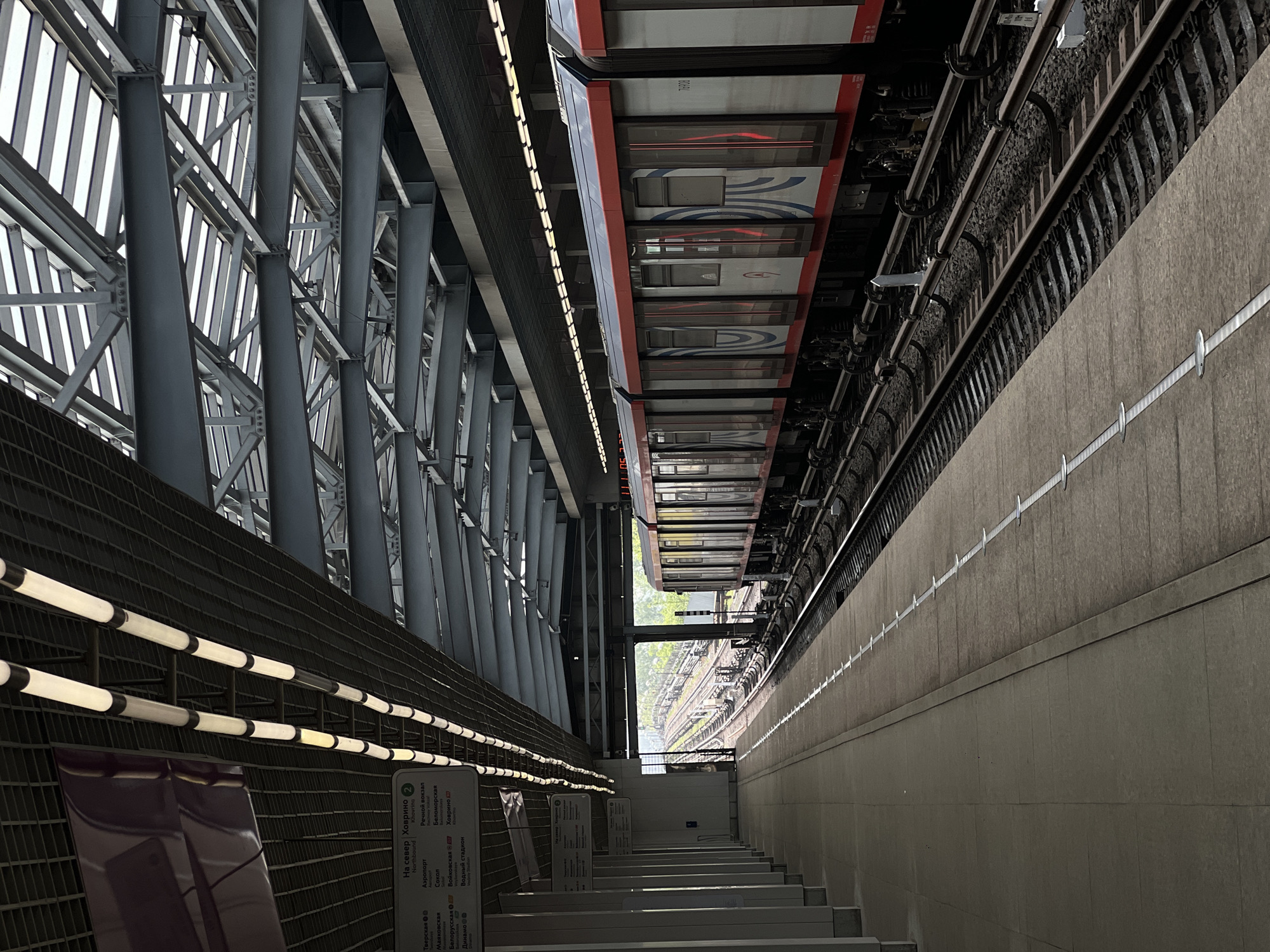The image depicts an underground metro station, possibly in an urban setting, featuring a departing train. The station has a distinctive architectural design, with a lattice-like structure of steel beams supporting the ceiling and a series of cylindrical pillars. The train, colored in red and white, is positioned above the platform, appearing to be pulling away from the station. The tracks and supporting infrastructure are visible above the platform edge.
To the left of the frame, a set of stairs leads down into the station, illuminated by horizontal rows of lights. Signs with Cyrillic and English text indicate the station's name and directions. The platform is constructed of a light-colored concrete material and is marked with white lines. The overall scene suggests a modern, functional transportation hub within an urban environment.