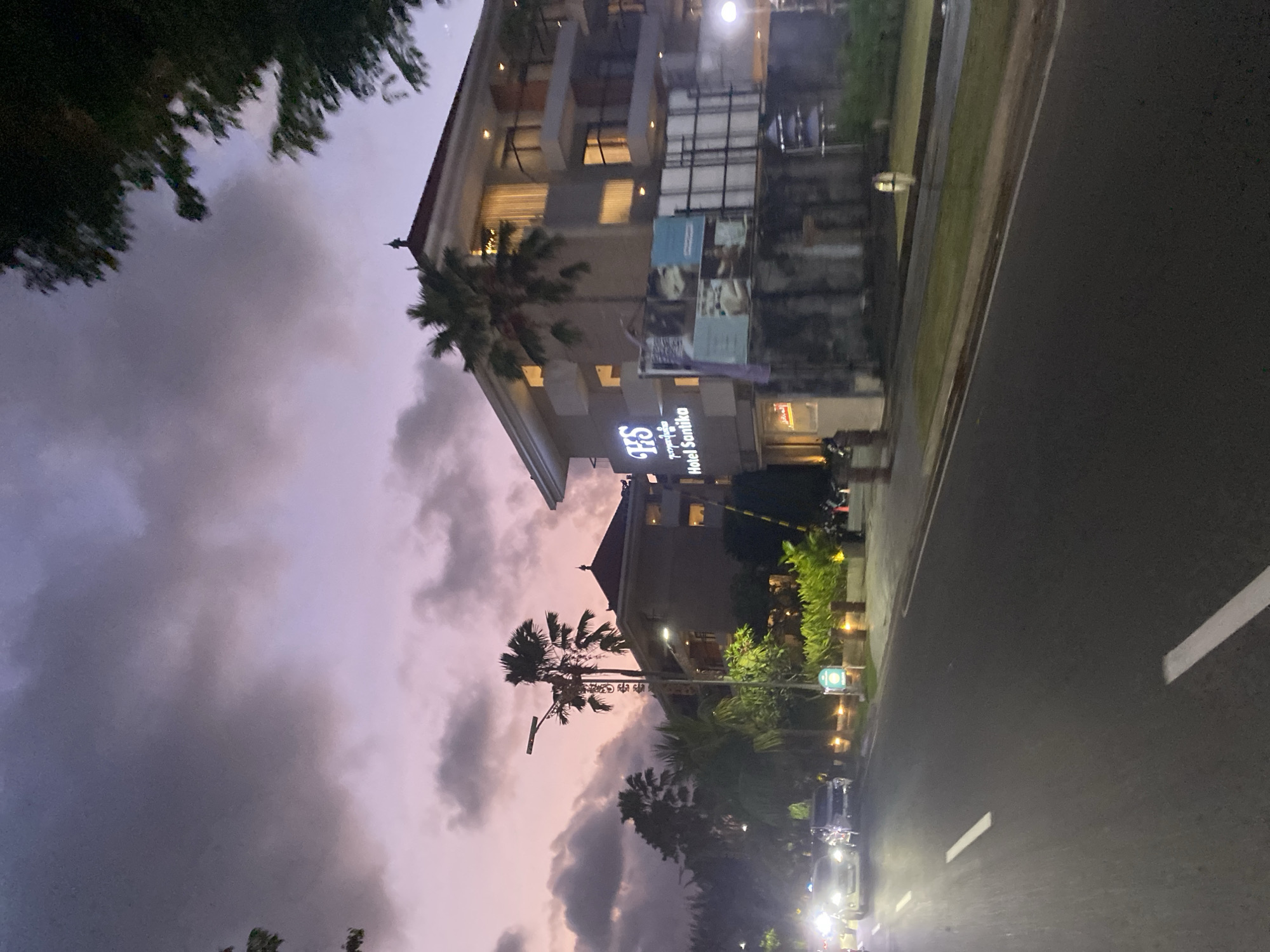 Captured in Kuta Selatan, Indonesia, the image depicts a street scene at dusk. A multi-story hotel with a sign reading "HS Hotel Sanika" stands prominently on the left side. Palm trees are interspersed along the building. To the left is a road, with cars visible at the bottom, and road markings. The sky is filled with purple and grey clouds. The image is taken from a low angle, looking up at the hotel and sky.