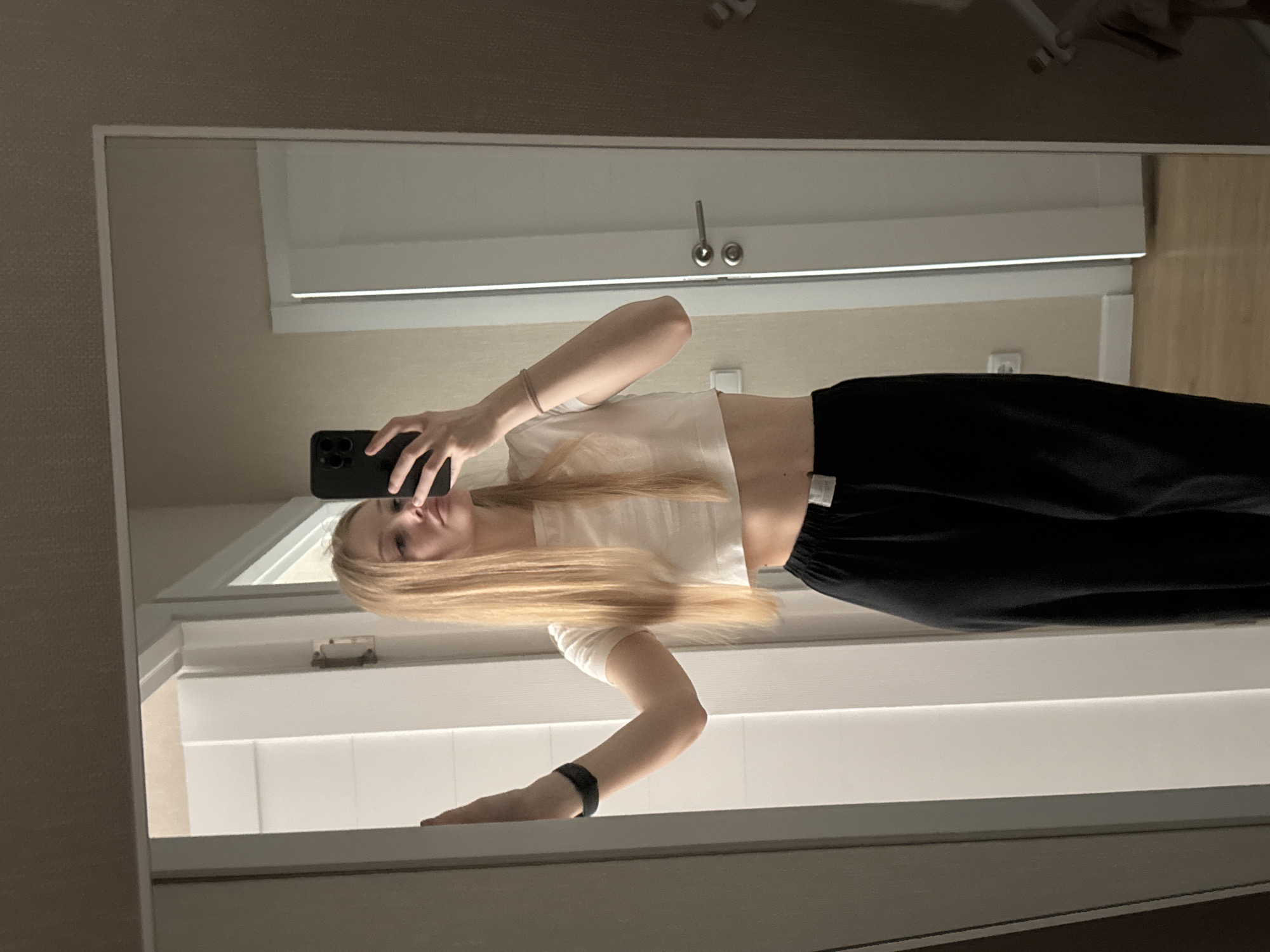 A young woman with long blonde hair is taking a mirror selfie in a dimly lit room. She is wearing a white crop top and black pants. She is standing in front of a full-length mirror, which reflects the room behind her. The room appears to be a bedroom or a dressing area, with a white door visible in the reflection. There is a light-colored wall and a wooden floor. The woman is holding her phone in her right hand, and her left arm is resting on the doorframe. A black fitness tracker is visible on her left wrist. A thin, light-colored bracelet is on her right wrist. There is no visible indication of the city of Perm, Russia.