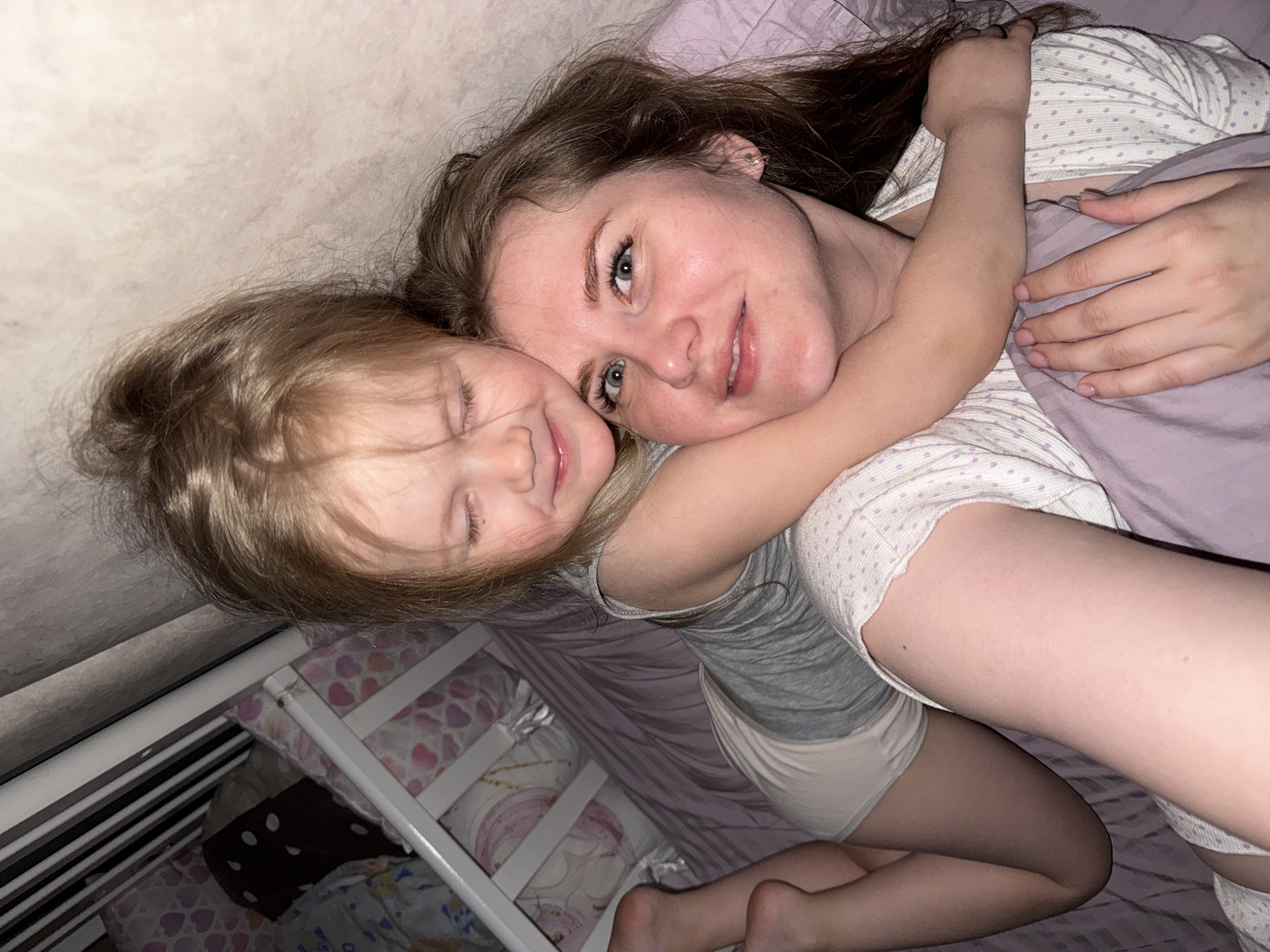 A woman and a young girl are lying on a bed. The woman has long brown hair and blue eyes, and she is wearing a white and lavender polka-dotted shirt. The girl has blonde hair and is wearing a gray shirt and white shorts. The girl has her arm around the woman. They are lying on a lavender bedspread. A white crib with patterned bedding is visible in the lower left corner of the image. The scene appears to be indoors, likely a bedroom. The location is specified as Gornyi (Dvurechenskoe S/P), Russia.
