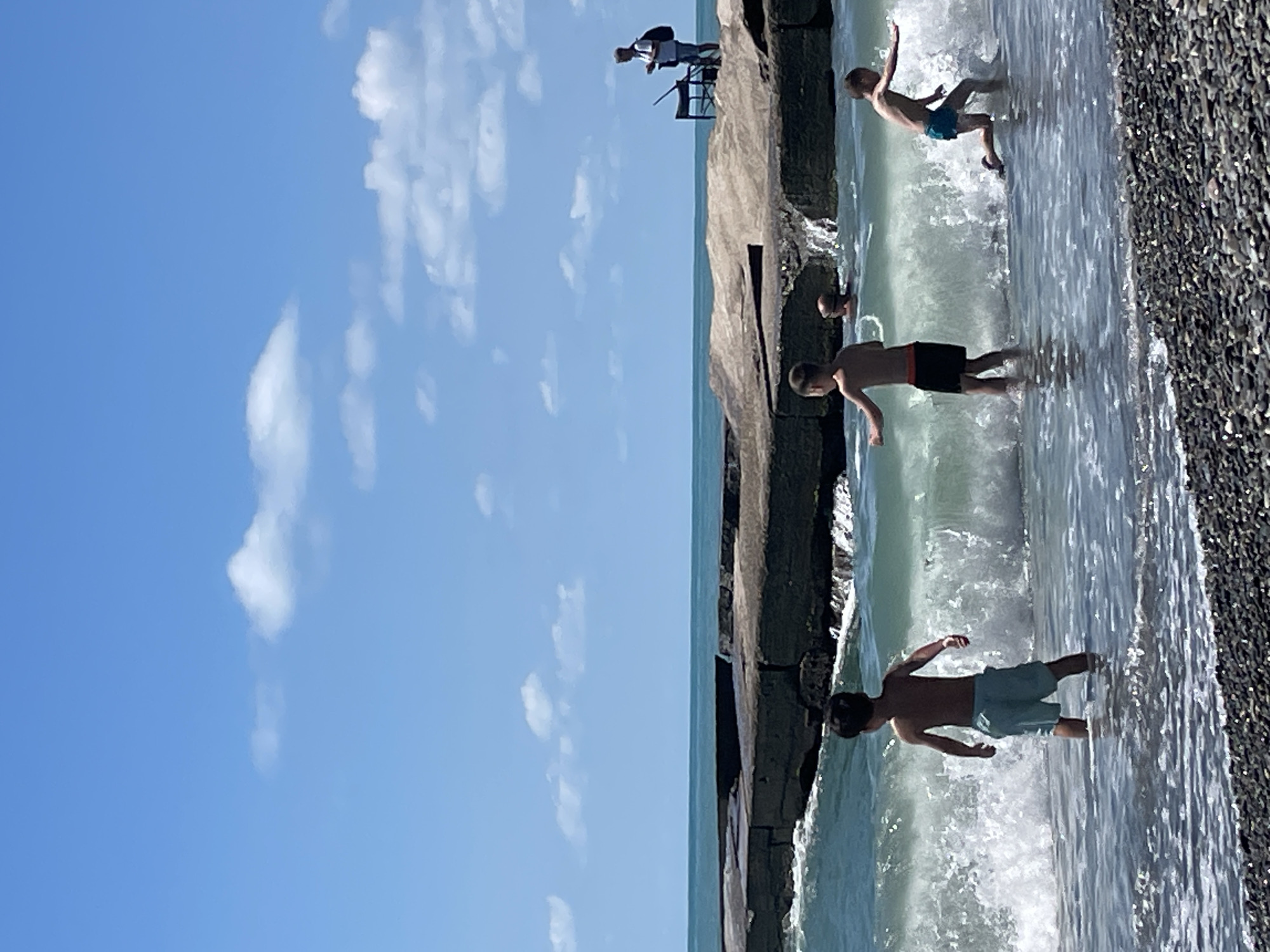 This image depicts a sunny day at the beach in Sochi, Russia. The foreground shows a pebbled shoreline leading into the Black Sea, where several children are playing in the shallow water. Waves crash onto the shore, creating white foam. In the mid-ground, a group of children are visible, one boy in light blue shorts stands facing away from the camera, partially submerged in the water. To his right, another boy in black shorts is also in the water, his arm outstretched. To the far right, a third boy in blue swim trunks is running into the waves. In the background, a large, dark, flat rock formation extends into the sea. On top of this rock formation, a man stands near a table with what appears to be a laptop, facing the sea. The sky above is a clear blue with scattered white clouds. The overall scene is one of leisure and recreation on a warm day.