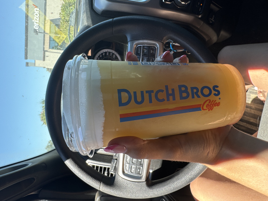 Inside a stationary vehicle in Granite Bay, California, United States, a person's left hand holds a large clear plastic Dutch Bros Coffee cup. The beverage is yellow-orange with a white foam topping. The hand exhibits a French manicure with pink and white polish. Visible vehicle components include a black steering wheel with integrated controls, a dashboard, and keys in the ignition. Through the windshield, a commercial building with 'Verizon' signage is visible, consistent with a parking lot at 5334 Sunrise Boulevard. The sky is clear and blue, with some distant trees also present.