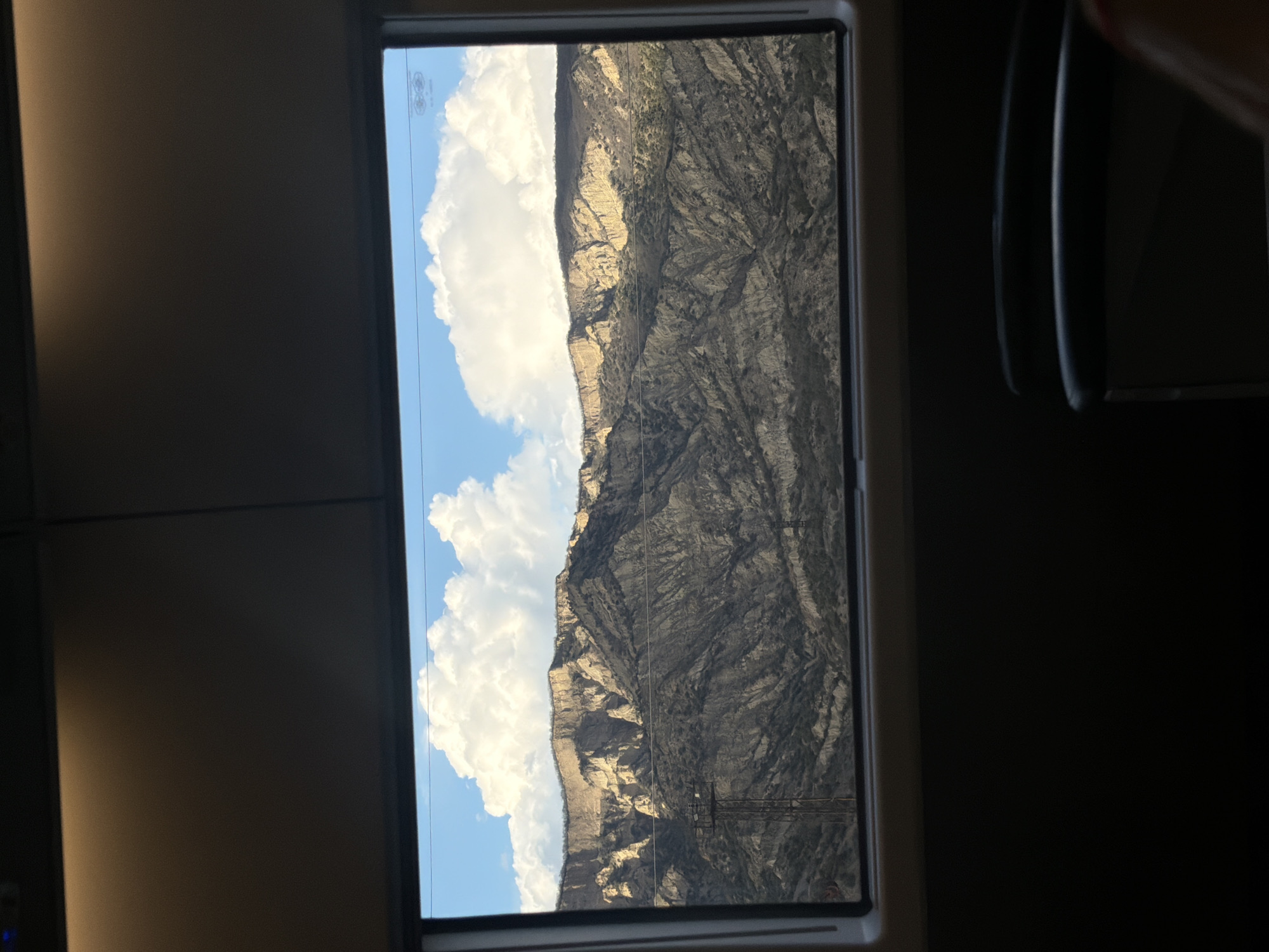 A view from a train window shows a mountainous landscape under a blue sky with cumulus clouds. Power lines are visible against the mountain. The train is dark inside, but a portion of a seat is visible on the right.